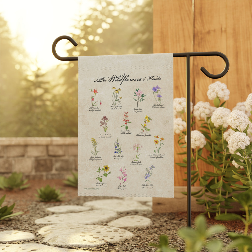 Decorative garden flag with illustrations of wildflowers on a beige background, attached to a black metal flagpole with a natural blurred background