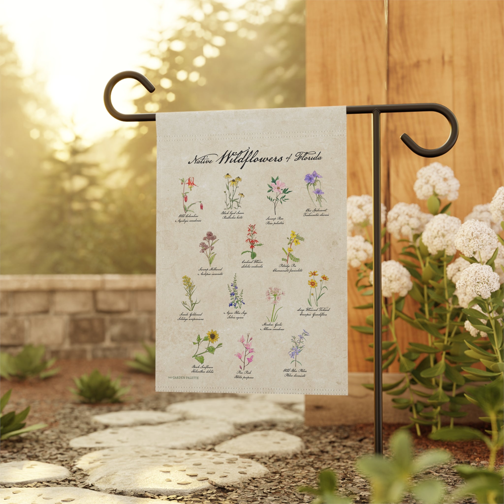 Decorative garden flag with illustrations of wildflowers on a beige background, attached to a black metal flagpole with a natural blurred background