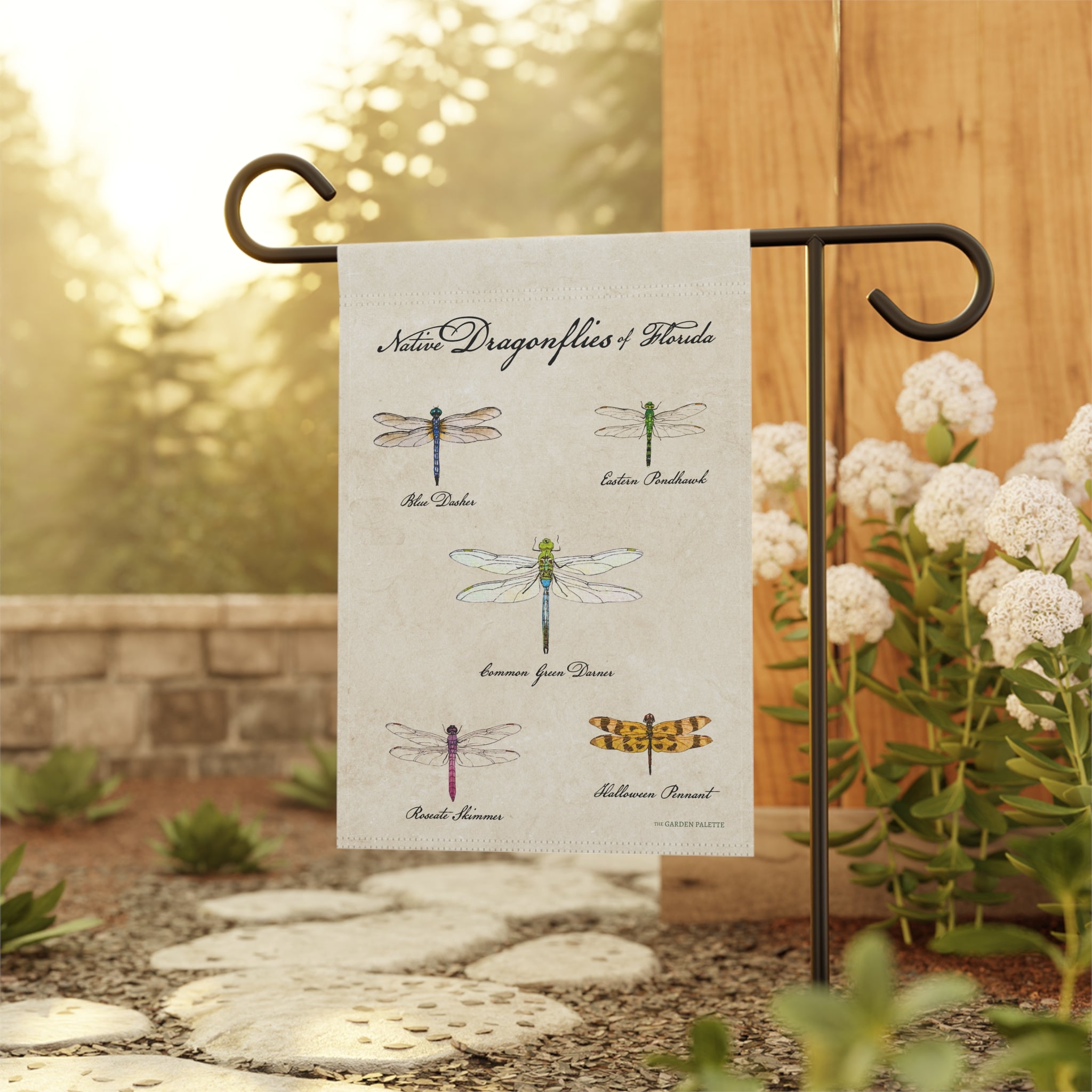 Decorative garden flag with illustrations of dragonflies on a beige background on a black pole with a blurred natural background