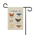 Garden flag with illustrations of butterflies and text on a beige background, held by a black metal pole.