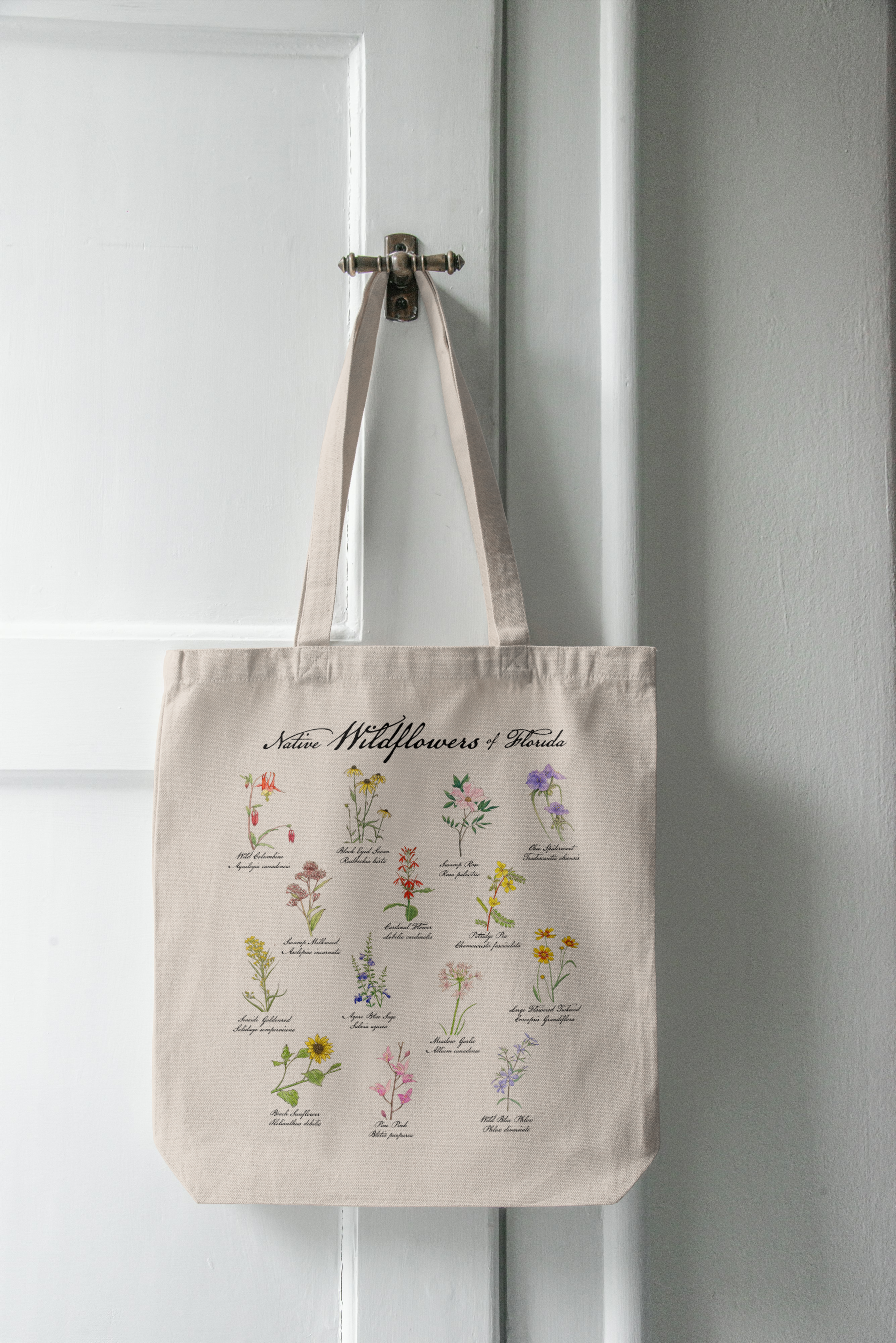 Beige tote bag with floral illustrations and text hanging on a white door.
