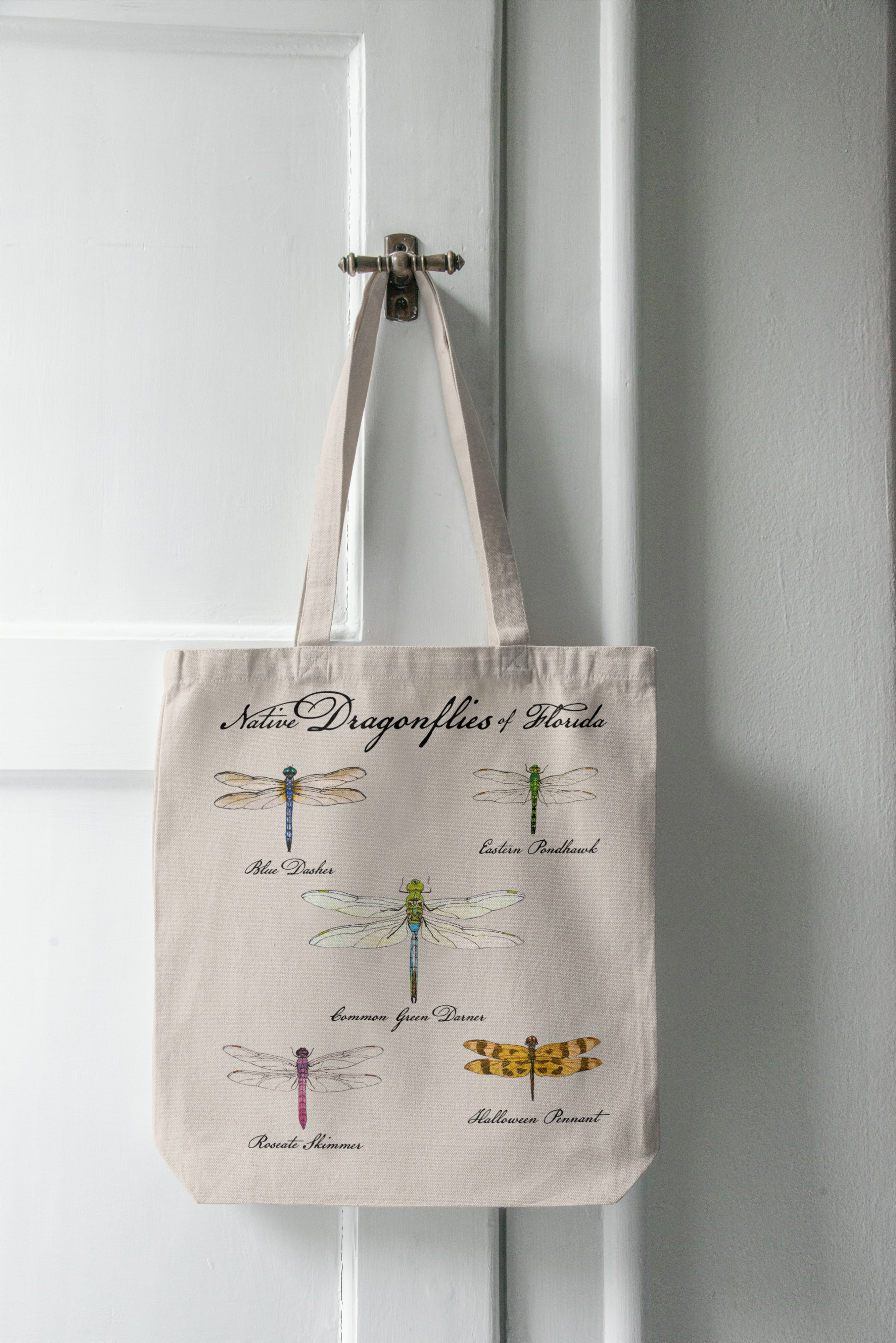 Tote bag with dragonfly illustrations hanging on a white door.