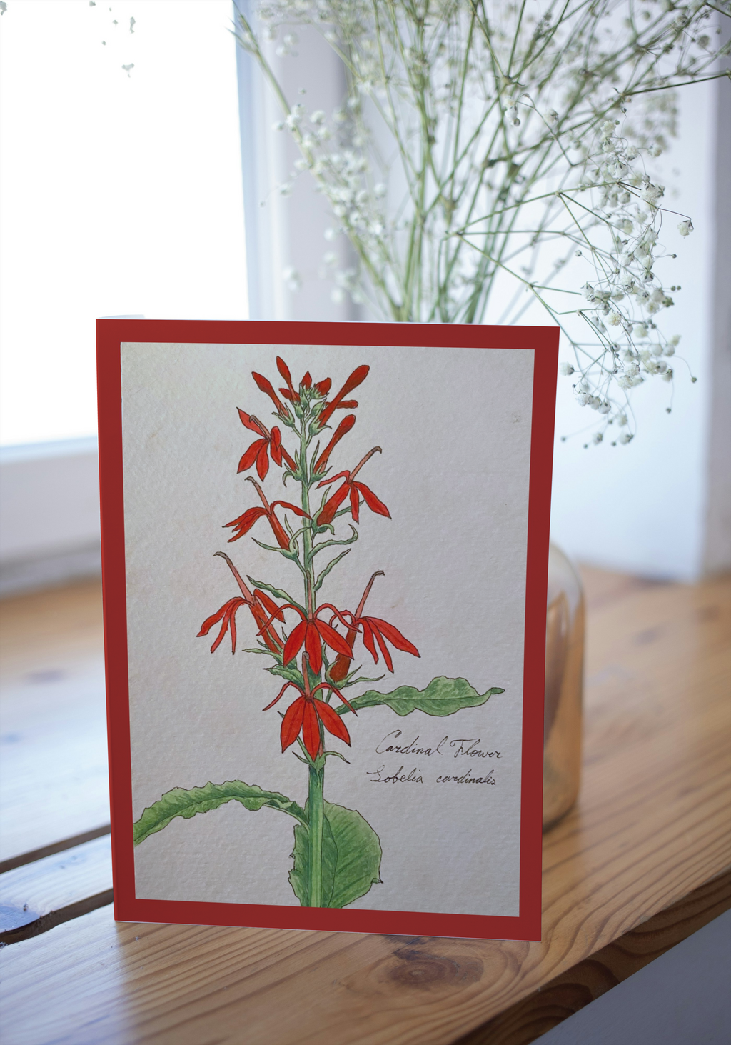 Card with red floral illustration on a wooden surface with flowers in the background