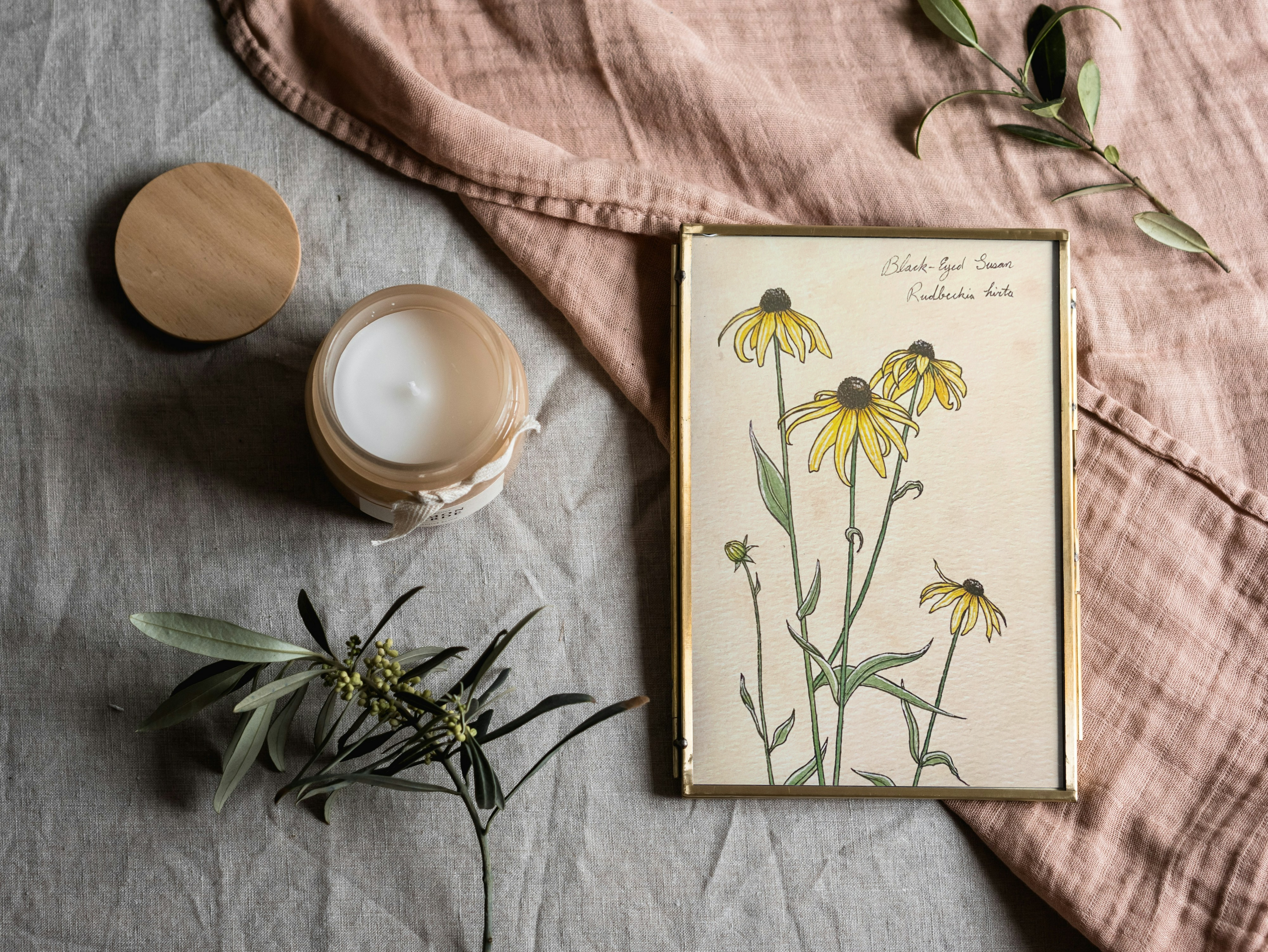 Framed botanical print of yellow flowers, candle, and greenery on a textured surface.