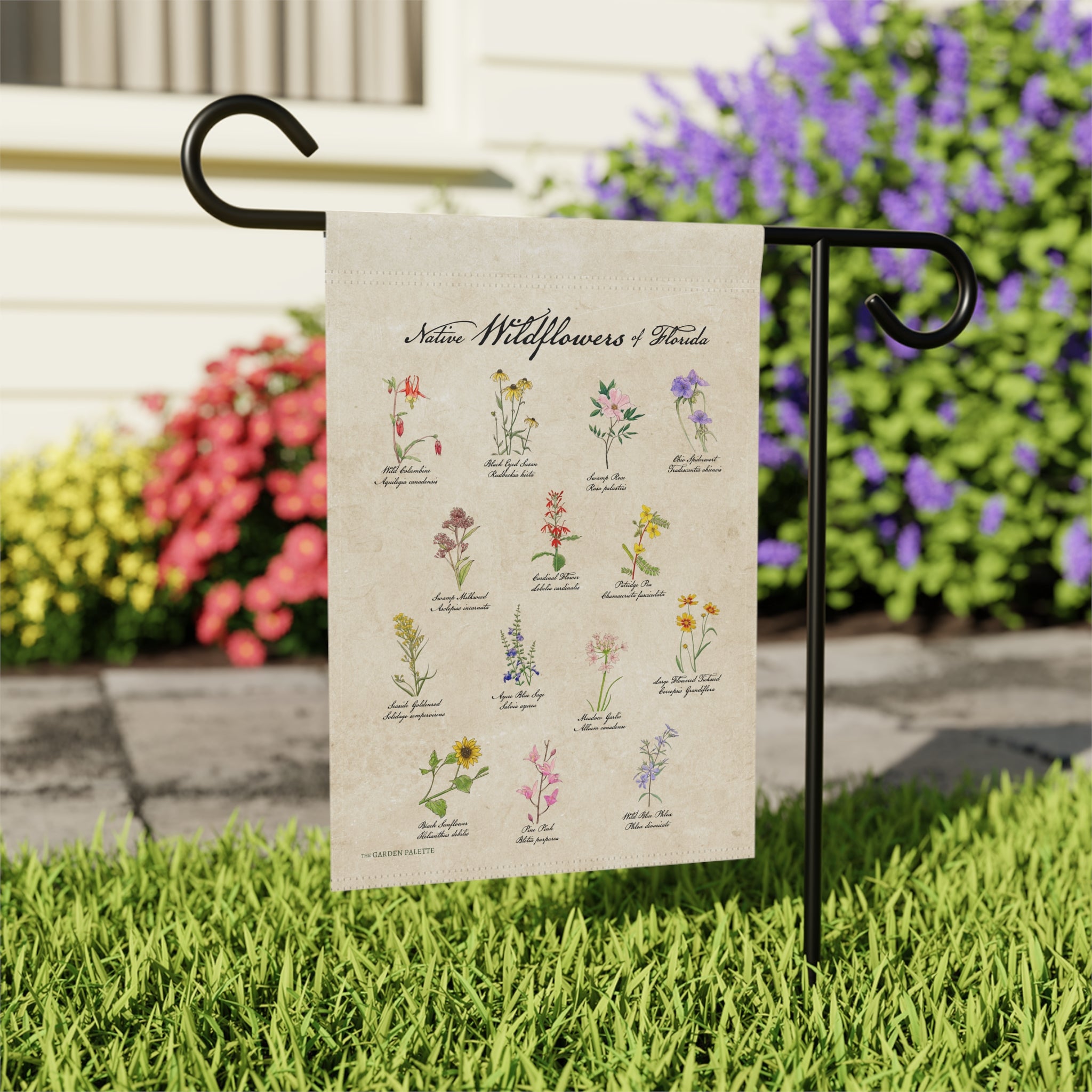 Decorative garden flag with illustrations of wildflowers on a beige background, attached to a black metal flagpole in a grassy lawn