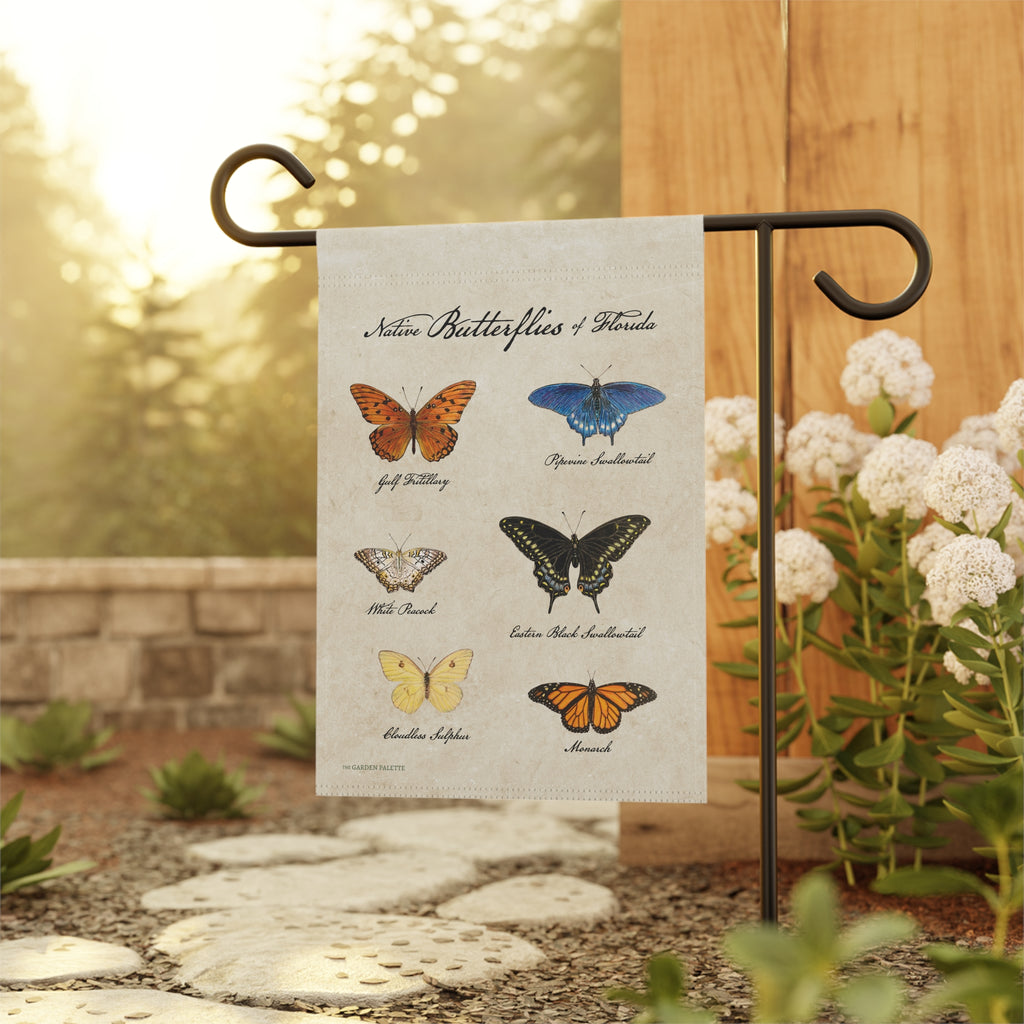 Garden flag with illustrations of butterflies and text on a beige background, held by a black metal pole with a natural blurred background