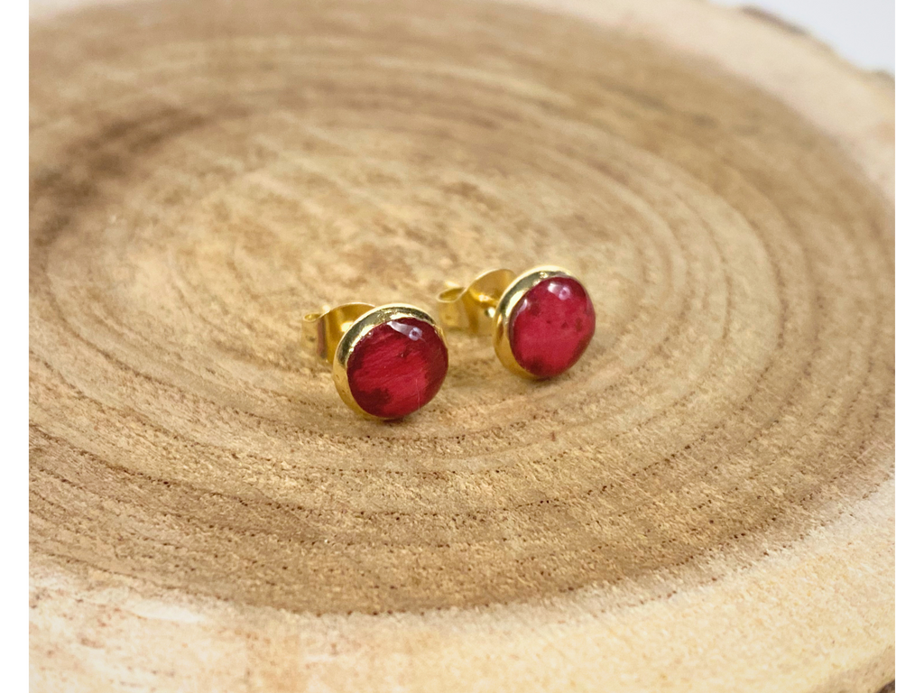 A pair of gold stud earrings containing deep pink flower petals, displayed a wooden slice background.