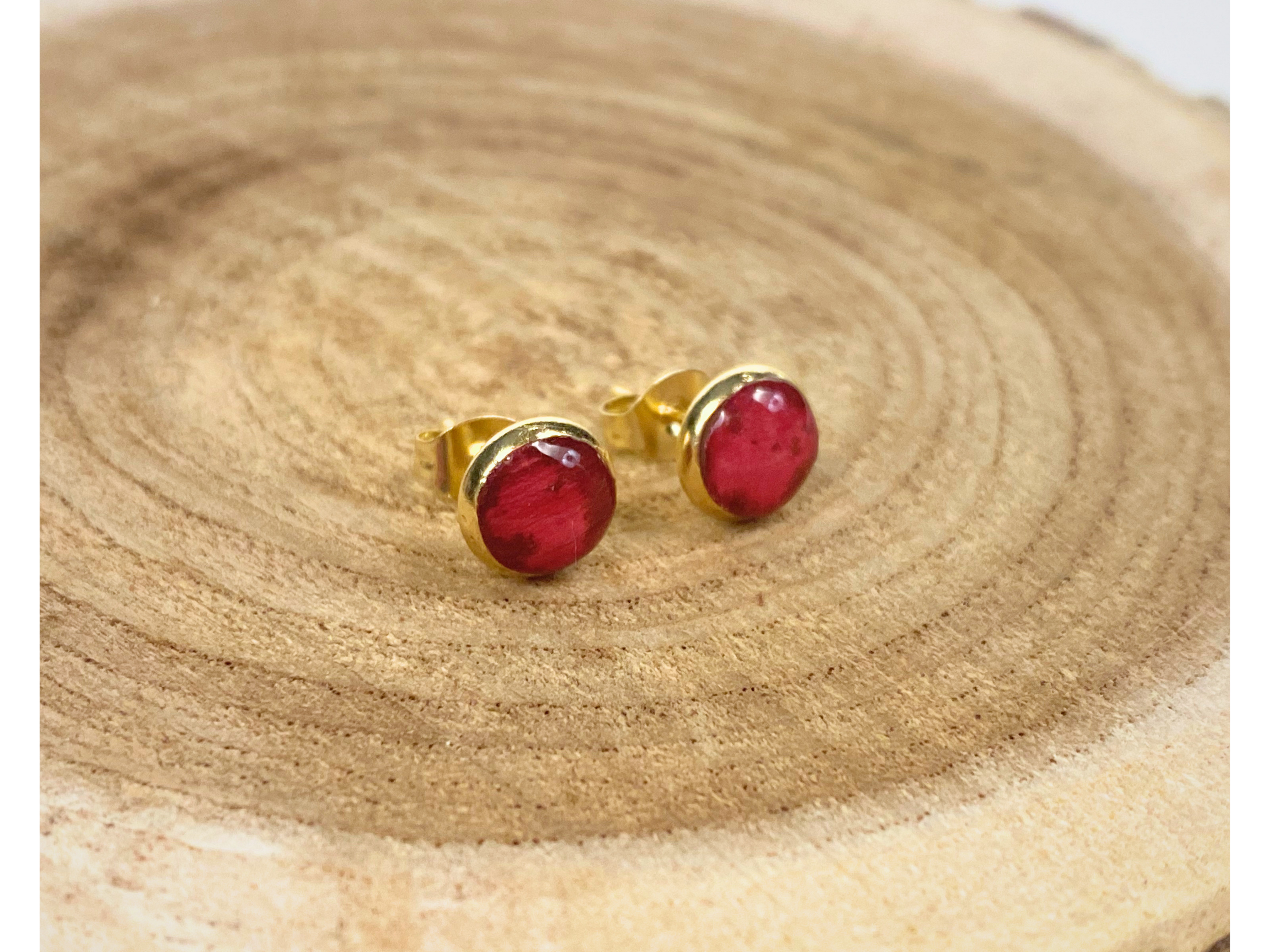 A pair of gold stud earrings containing deep pink flower petals, displayed a wooden slice background.
