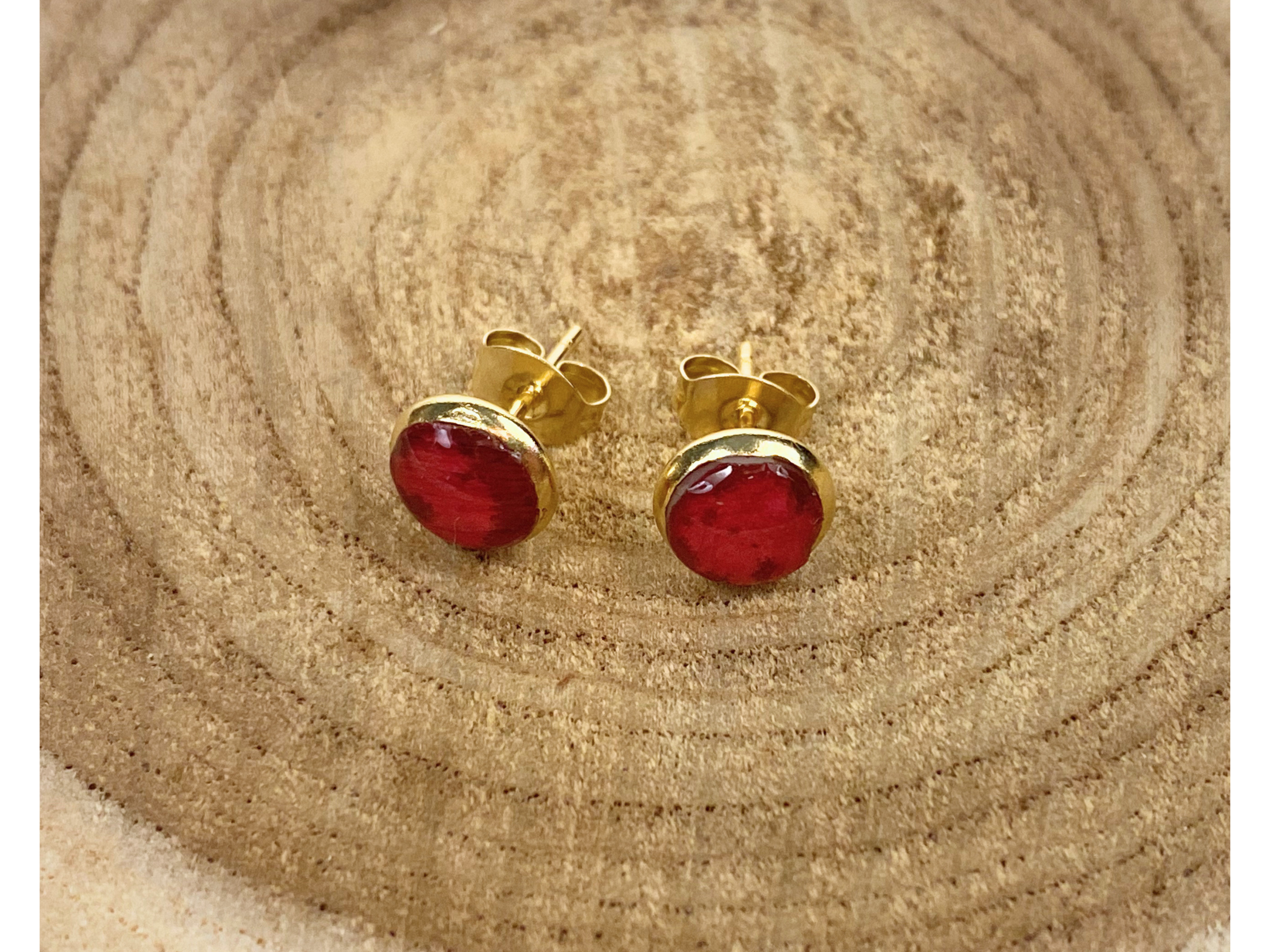 A pair of gold stud earrings containing deep pink flower petals, displayed a wooden slice background.