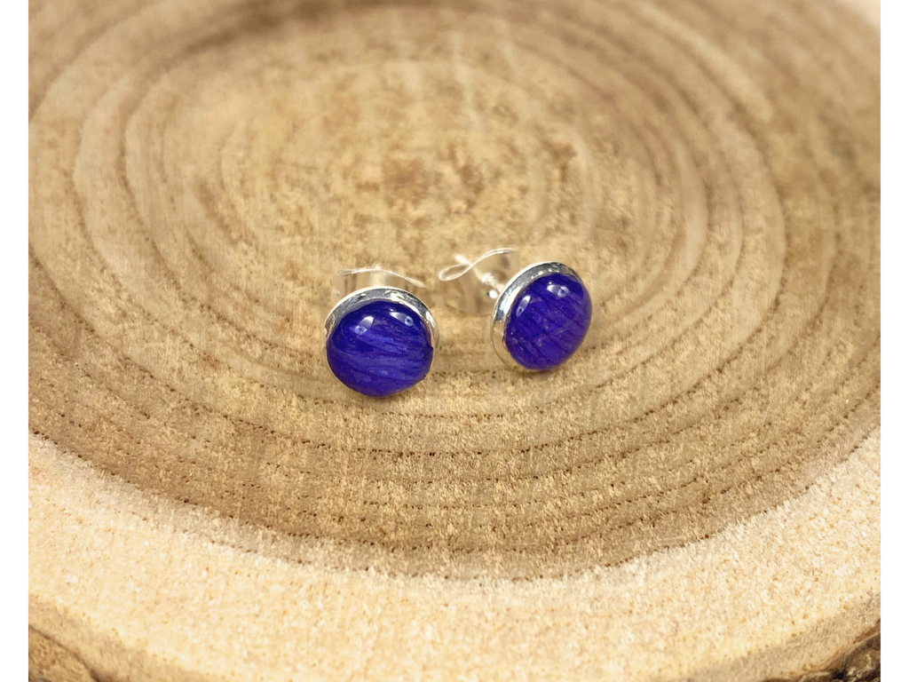 A pair of round silver stud earrings containing deep blue flower petals, displayed on a rustic wood slice background.