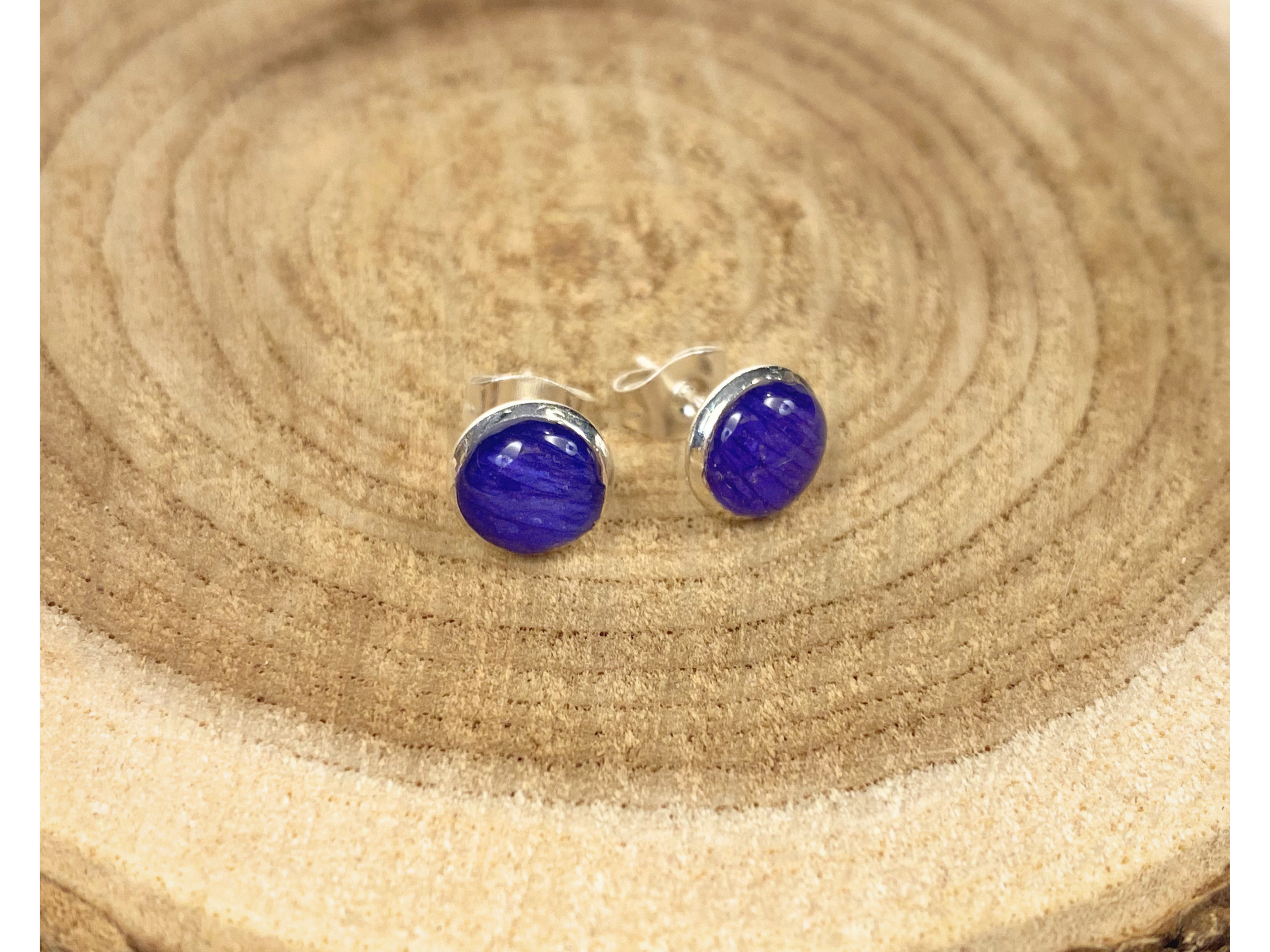 A pair of round silver stud earrings containing deep blue flower petals, displayed on a rustic wood slice background.