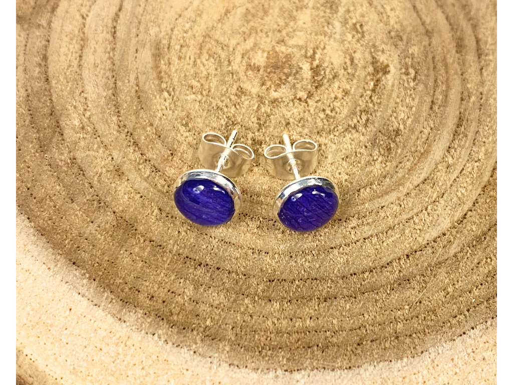 A pair of round silver stud earrings containing deep blue flower petals, displayed on a rustic wood slice background.
