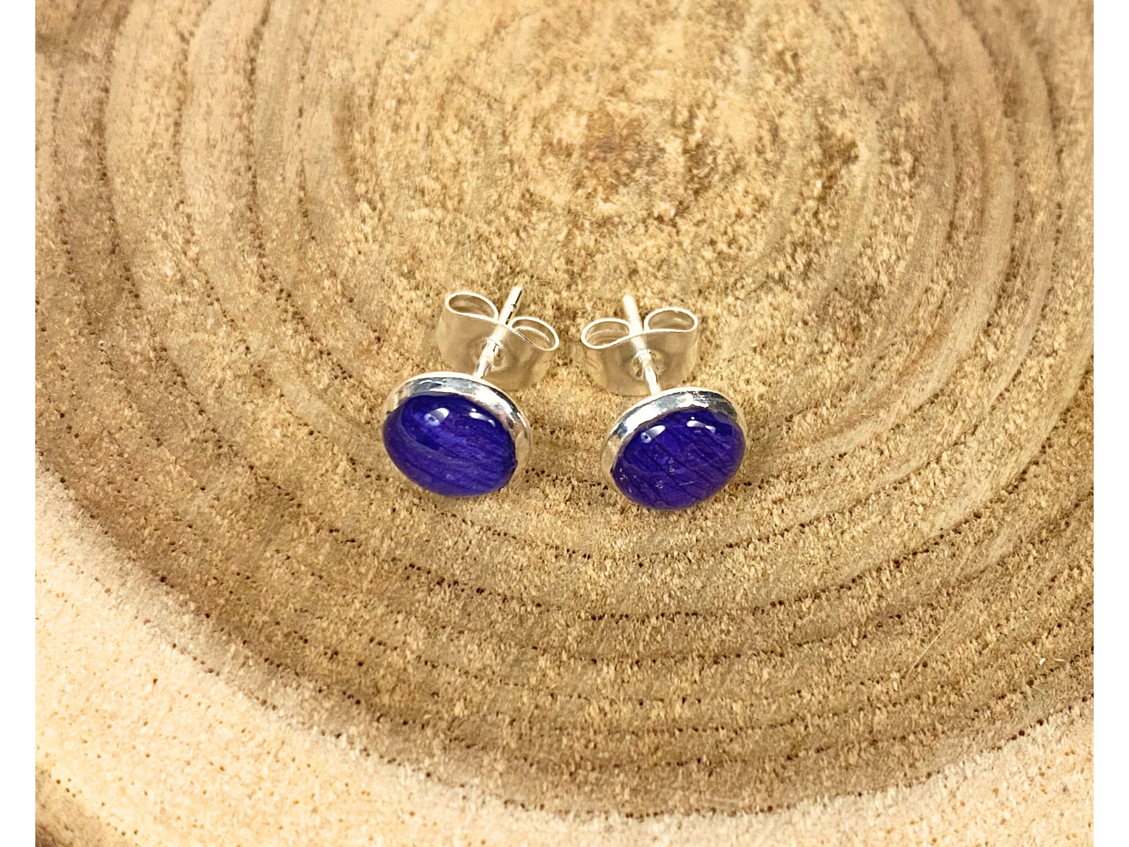 A pair of round silver stud earrings containing deep blue flower petals, displayed on a rustic wood slice background.