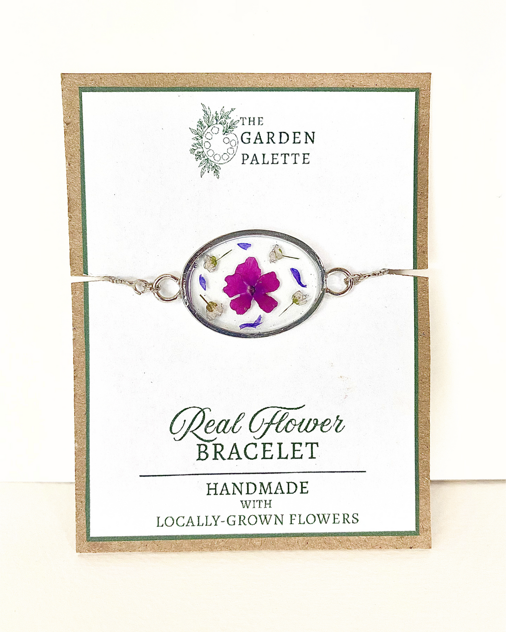 A silver oval pendant bracelet featuring a preserved purple flower and small botanical elements inside clear resin, displayed on a branded card that reads 'The Garden Palette' and 'Real Flower Bracelet'.