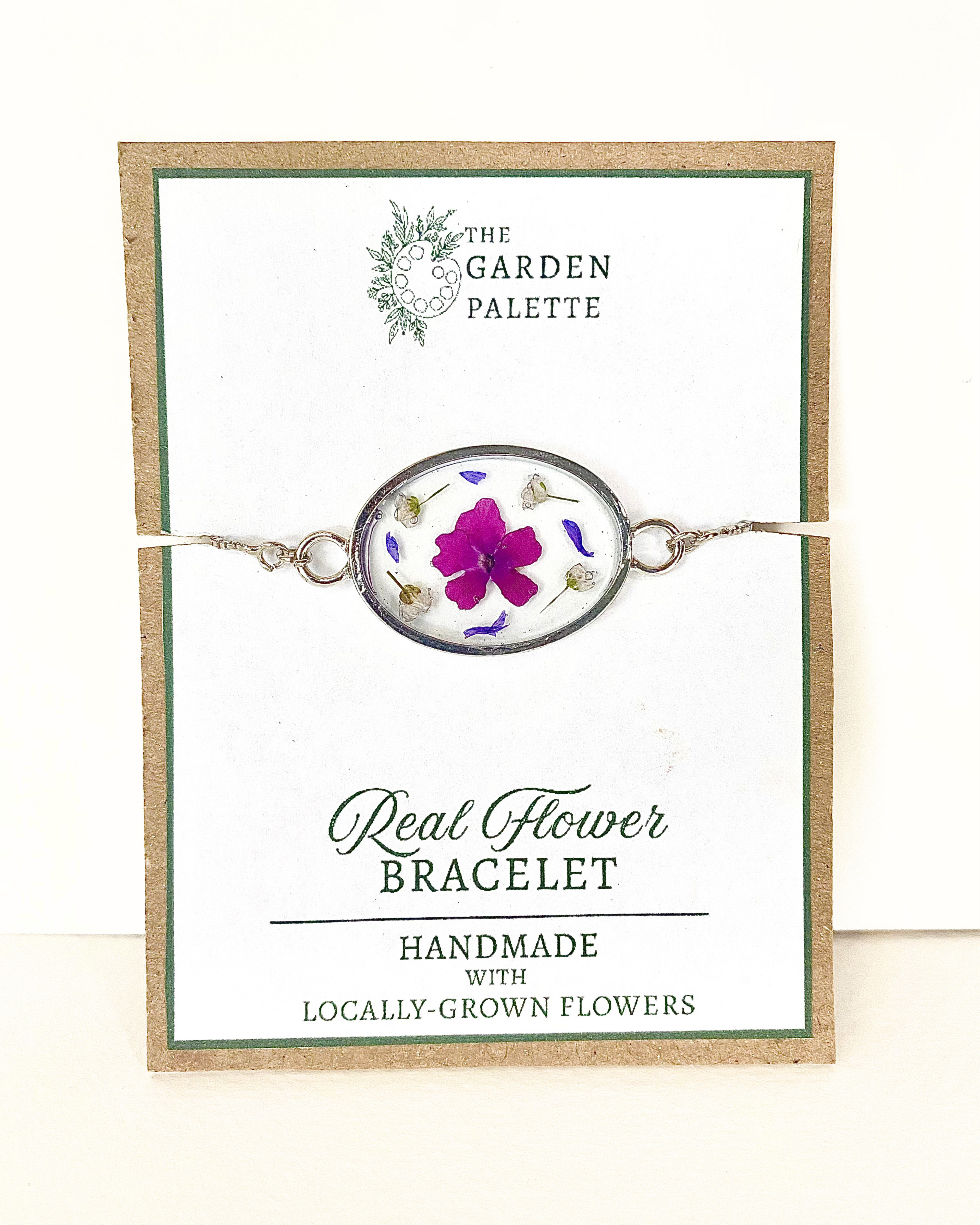 A silver oval pendant bracelet featuring a preserved purple flower and small botanical elements inside clear resin, displayed on a branded card that reads 'The Garden Palette' and 'Real Flower Bracelet'.