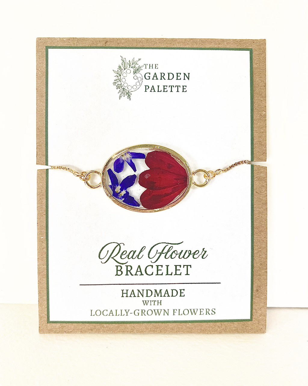 A handmade oval flower bracelet featuring red and blue real flowers in a gold-toned frame, displayed on its branded card.
