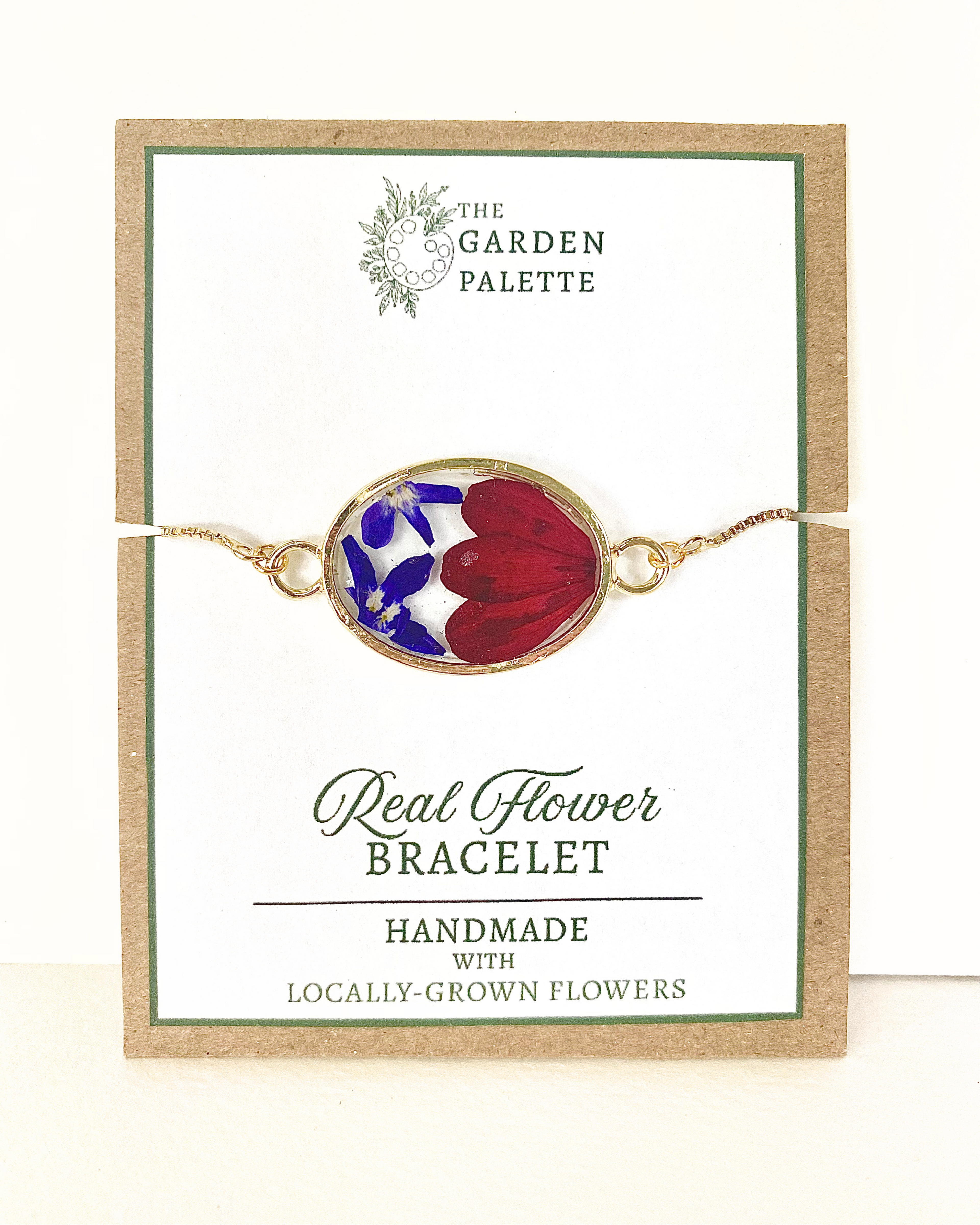 A handmade oval flower bracelet featuring red and blue real flowers in a gold-toned frame, displayed on its branded card.