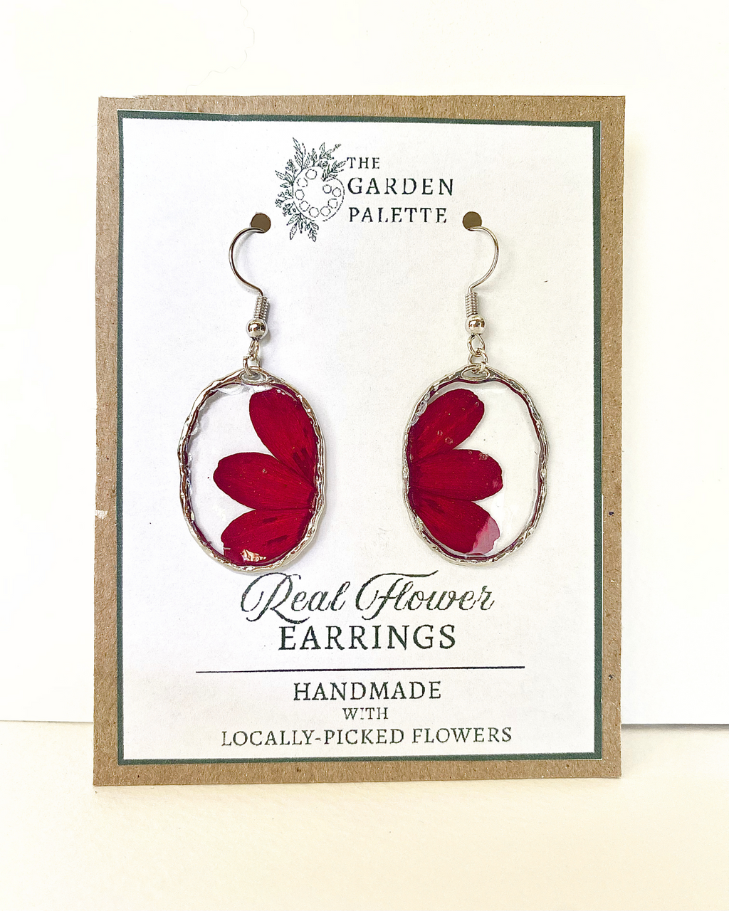 A pair of handmade oval dangle earrings with deep red pressed flowers in silver-toned frames, displayed on a branded card that reads 'The Garden Palette' and 'Real Flower Earrings'.
