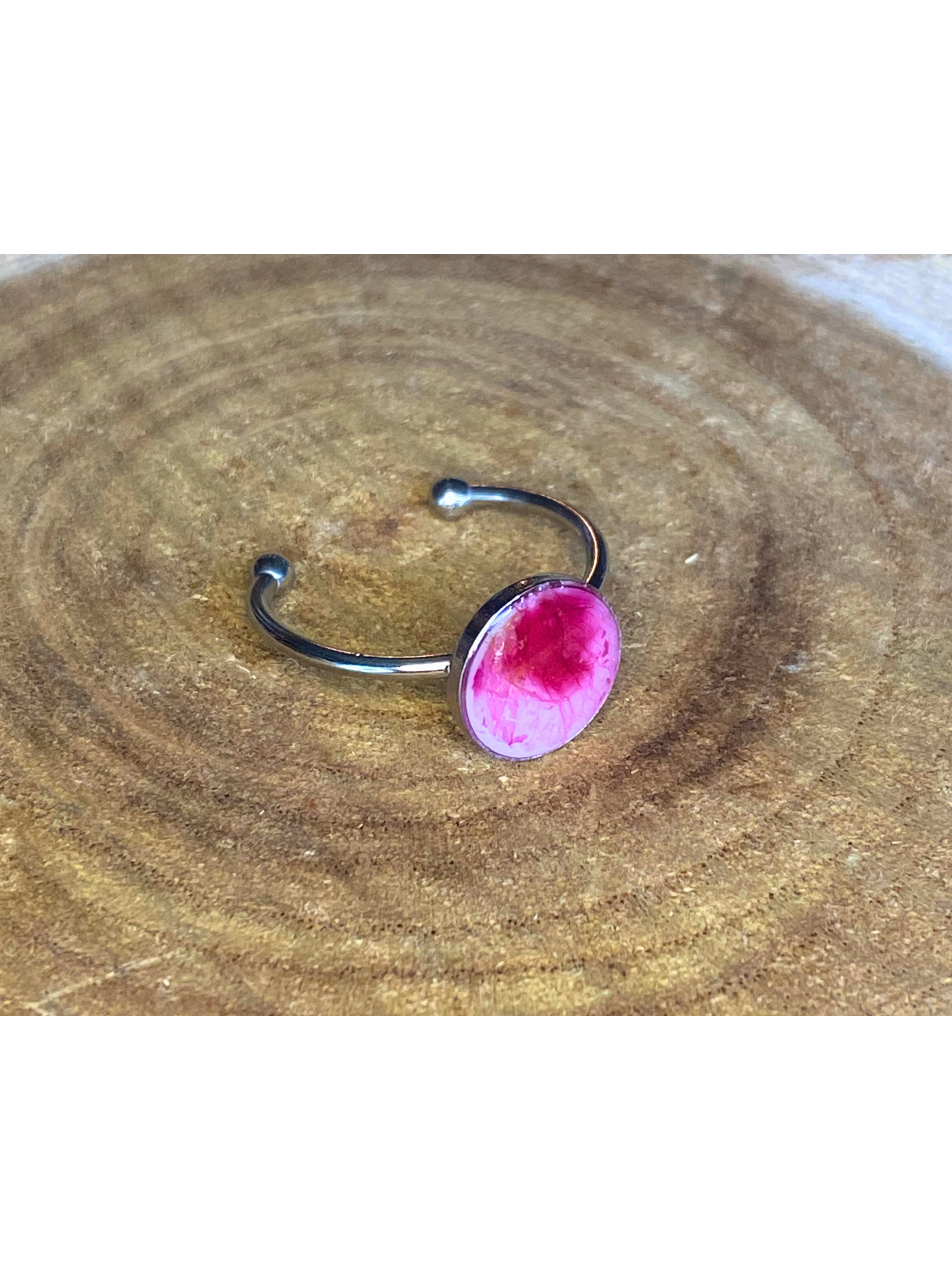 A silver-toned ring with a round bezel setting containing a pink petal, displayed on a rustic wood background. 