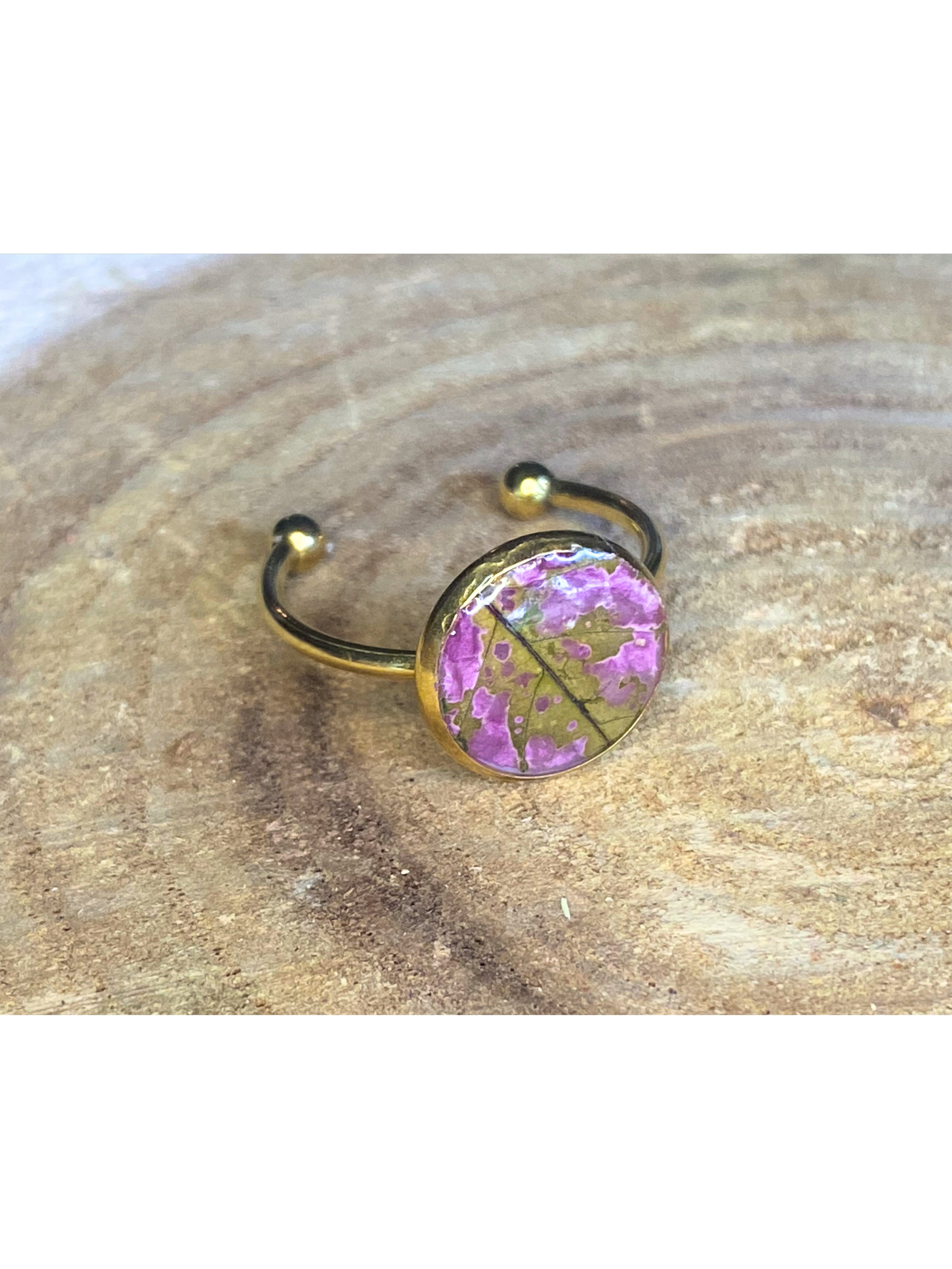 A silver-toned ring with a round bezel setting containing a pink and green leaf, displayed on a rustic wood background.