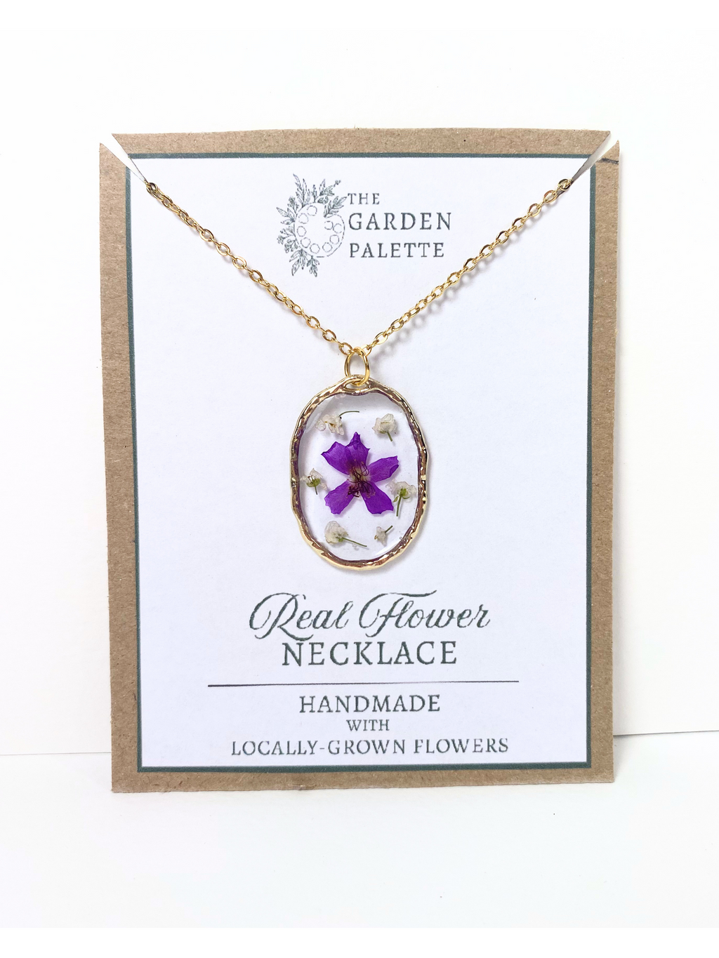A gold-tone oval pendant necklace with a purple flower and dried botanicals inside clear resin, displayed on a branded white card with a kraft paper border. The card reads 'The Garden Palette' and 'Real Flower Necklace'.