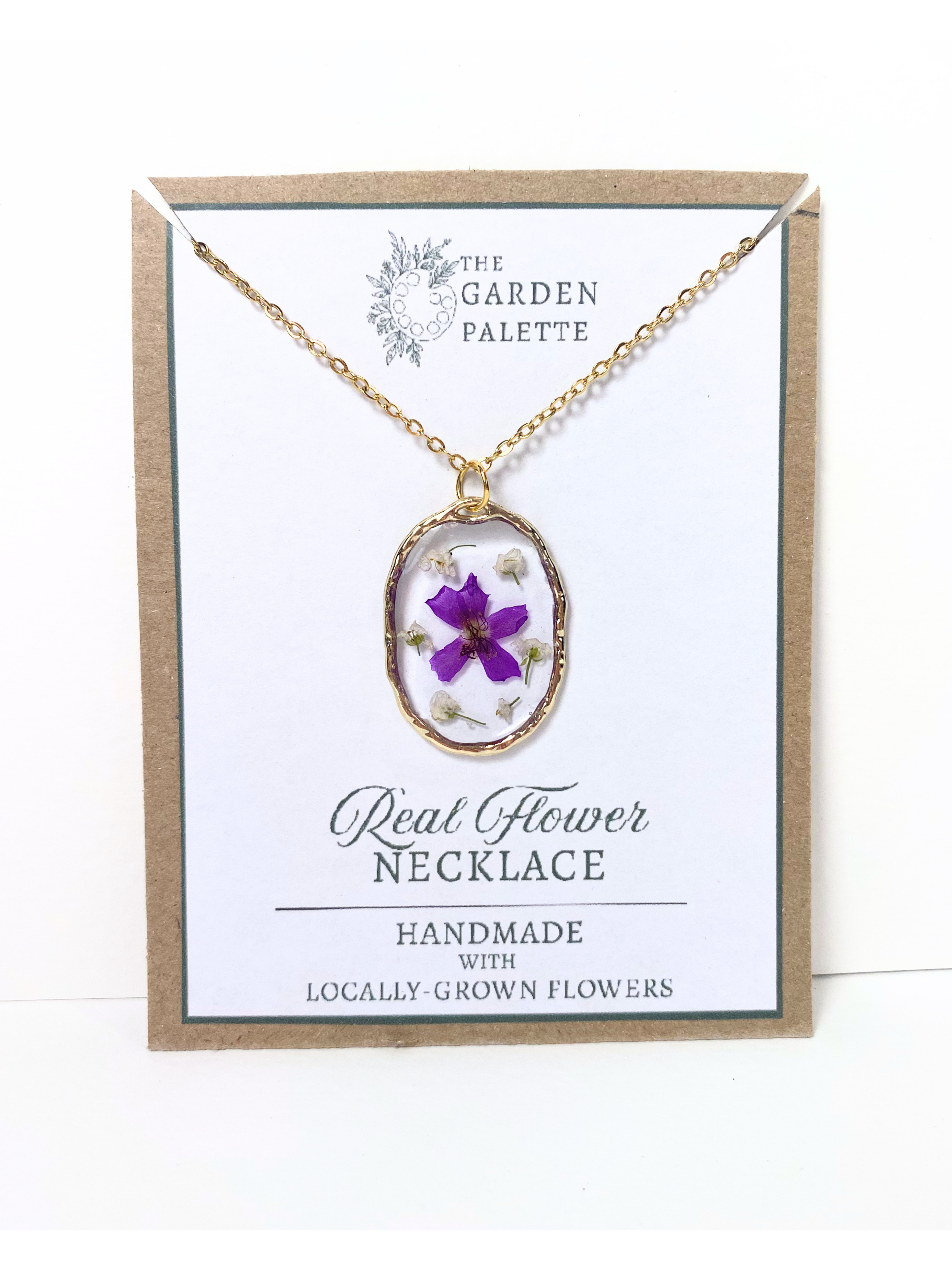 A gold-tone oval pendant necklace with a purple flower and dried botanicals inside clear resin, displayed on a branded white card with a kraft paper border. The card reads 'The Garden Palette' and 'Real Flower Necklace'.