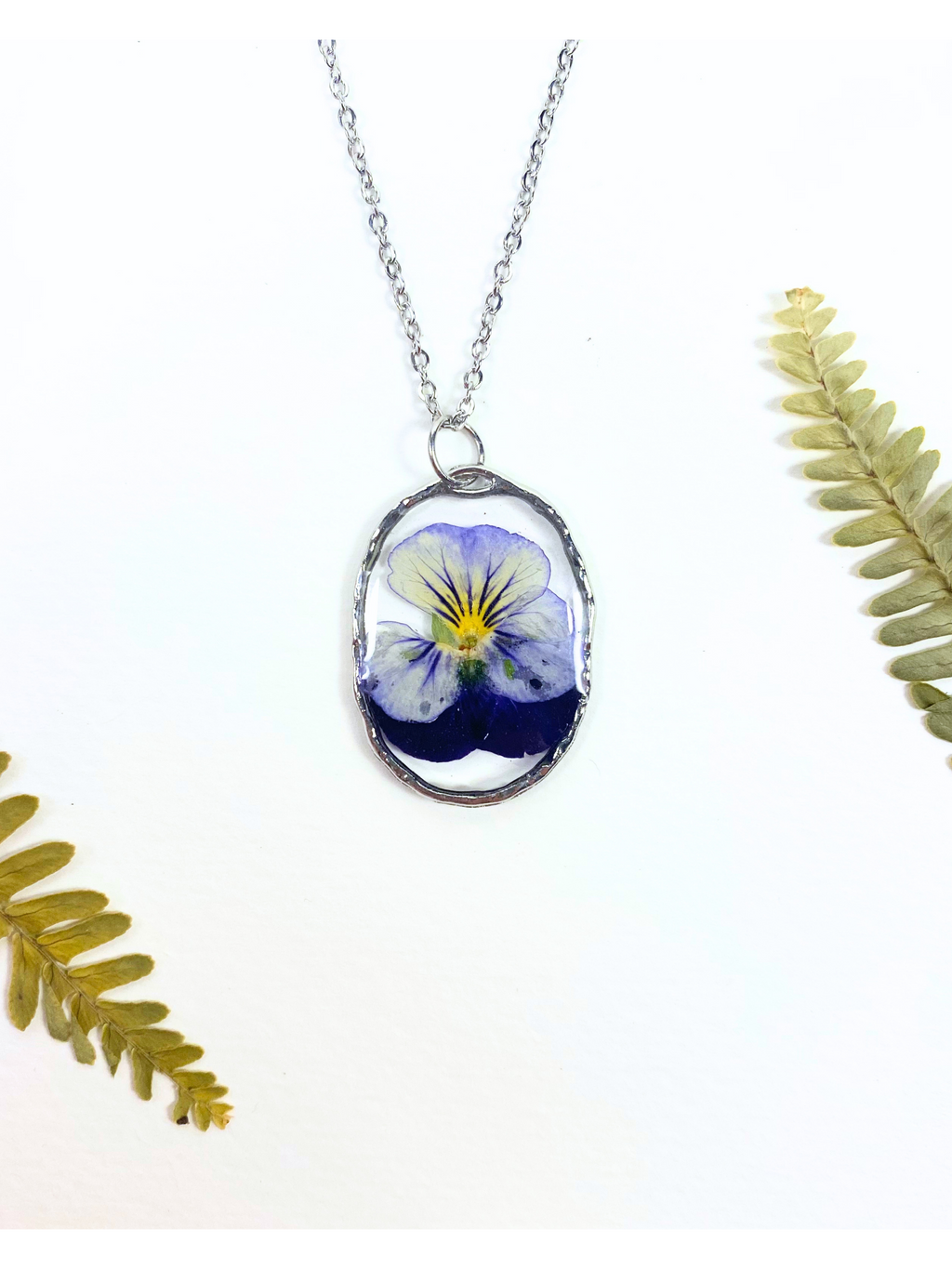 A silver oval pendant necklace featuring a preserved purple and yellow pansy flower under clear resin, displayed on a white background with decorative green fern leaves.