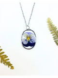 A silver oval pendant necklace featuring a preserved purple and yellow pansy flower under clear resin, displayed on a white background with decorative green fern leaves.