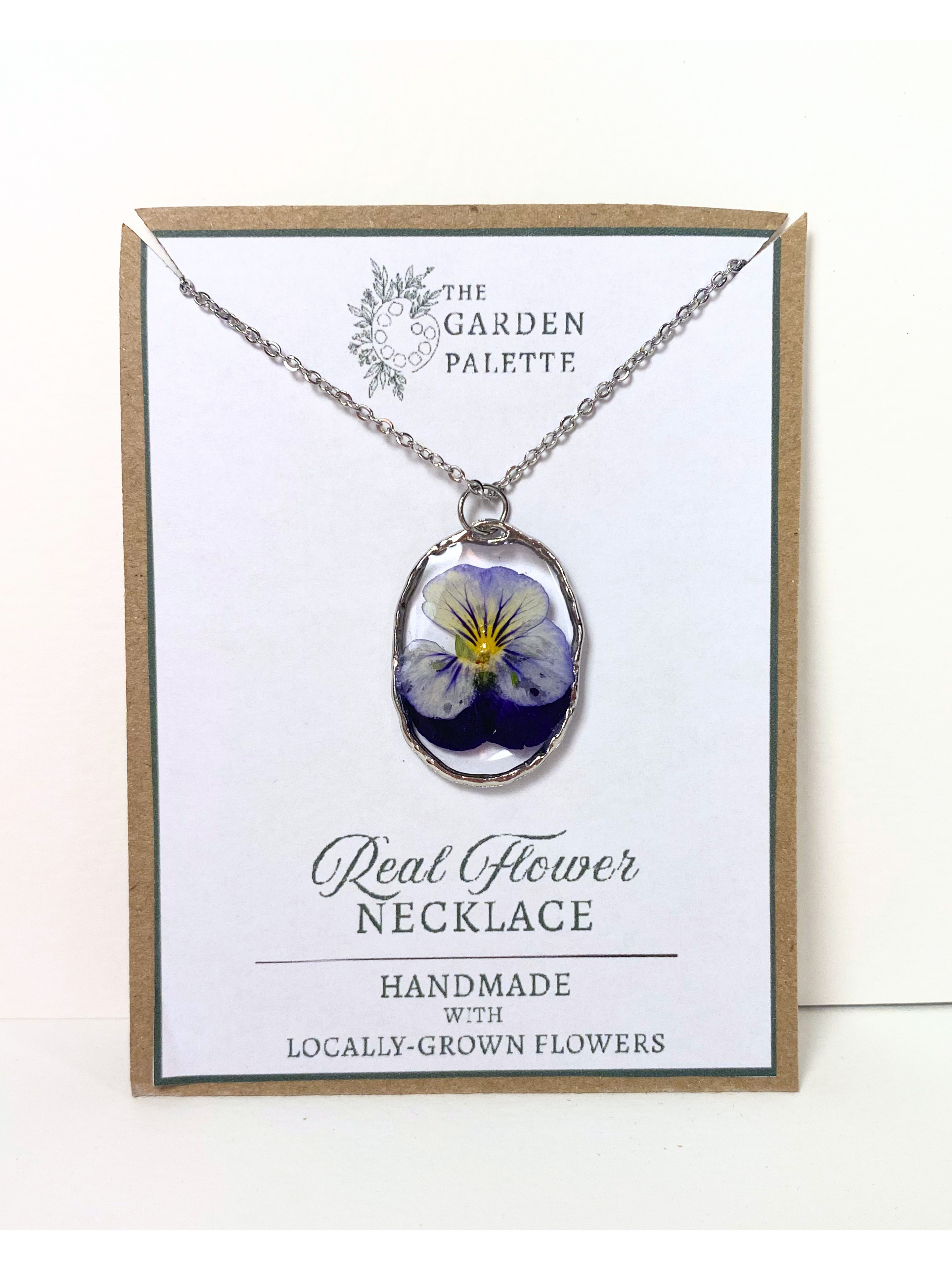 A handmade oval pendant necklace featuring a real purple and yellow pansy flower, displayed on a white card with a kraft paper border. The card is branded 'The Garden Palette' and reads 'Real Flower Necklace'.