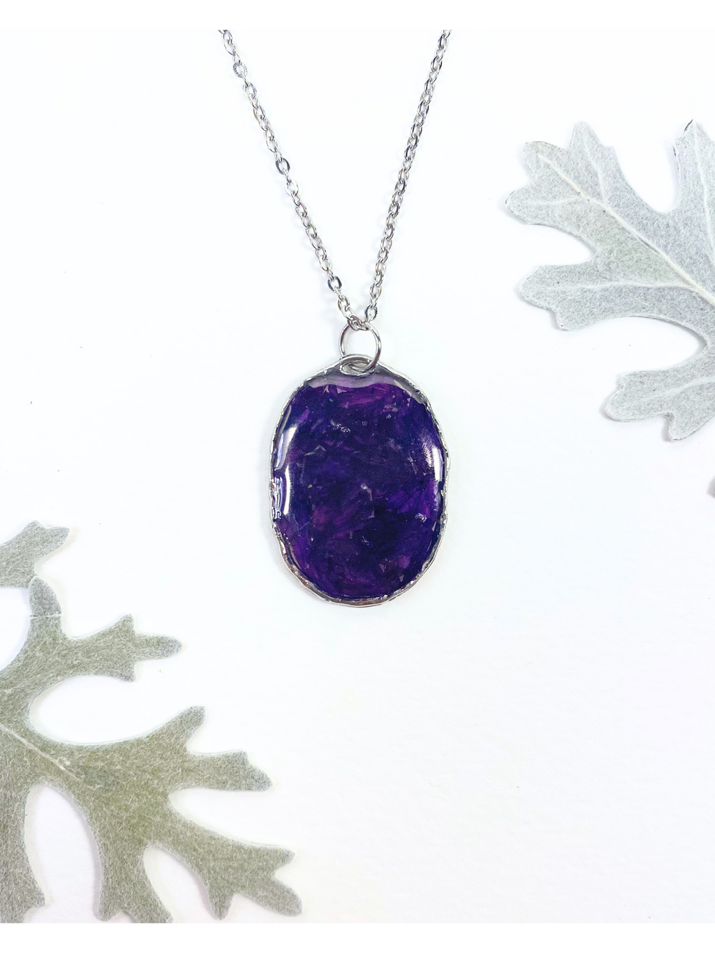 An oval pendant necklace containing deep purple flower petals with a silver chain, displayed on a white background with decorative leaves in the corners.