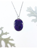 An oval pendant necklace containing deep purple flower petals with a silver chain, displayed on a white background with decorative leaves in the corners.