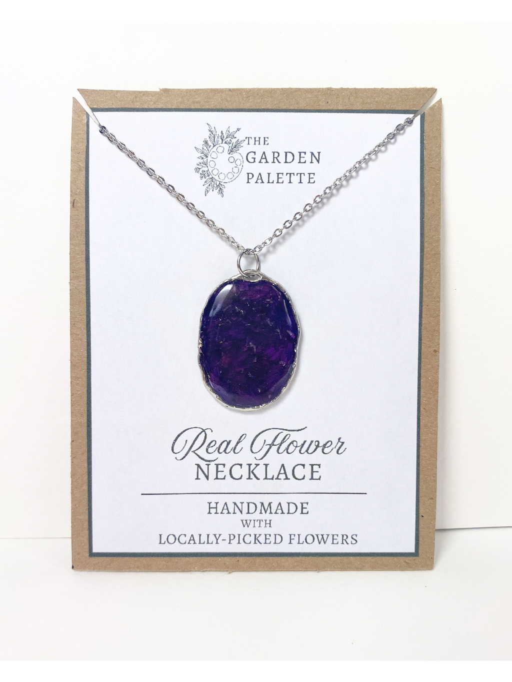A handmade oval pendant necklace containing deep purple flower petals on a silver chain, displayed on a white card with a kraft paper border. The card is branded 'The Garden Palette' and reads 'Real Flower Necklace'.