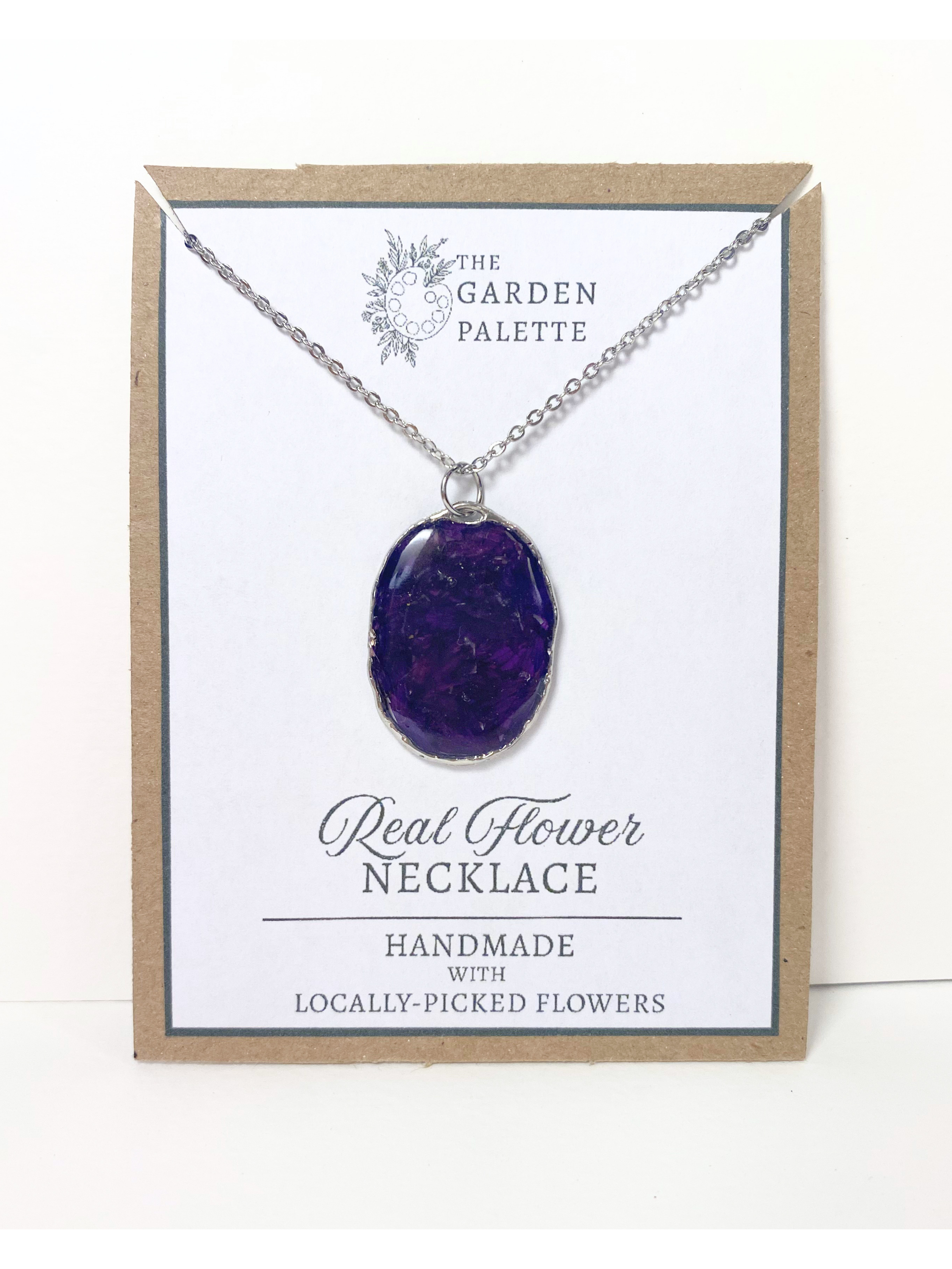 A handmade oval pendant necklace containing deep purple flower petals on a silver chain, displayed on a white card with a kraft paper border. The card is branded 'The Garden Palette' and reads 'Real Flower Necklace'.