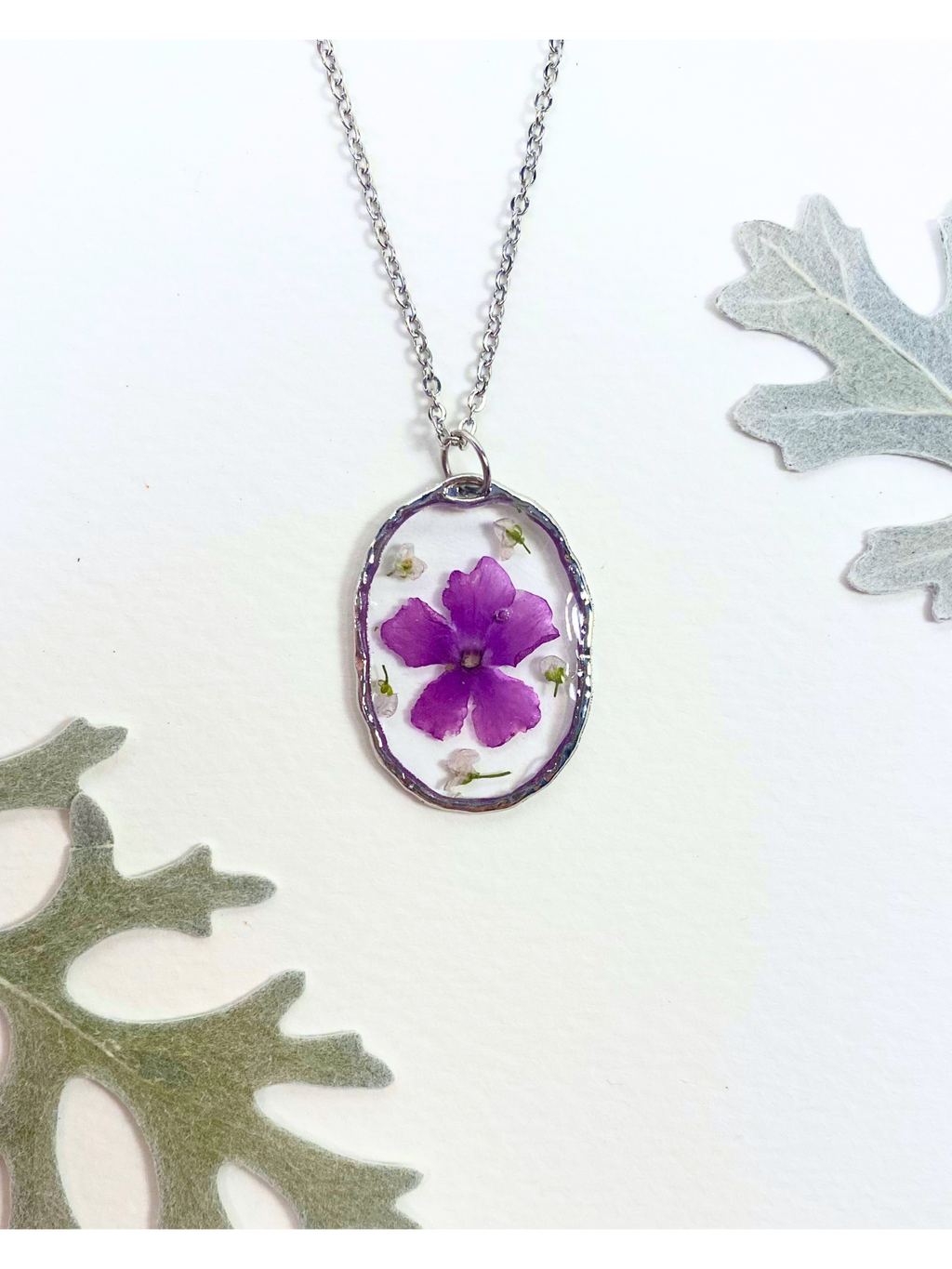 A silver oval pendant necklace featuring a preserved purple flower and small white flowers, displayed on a white background with decorative leaves.