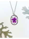 A silver oval pendant necklace featuring a preserved purple flower and small white flowers, displayed on a white background with decorative leaves.