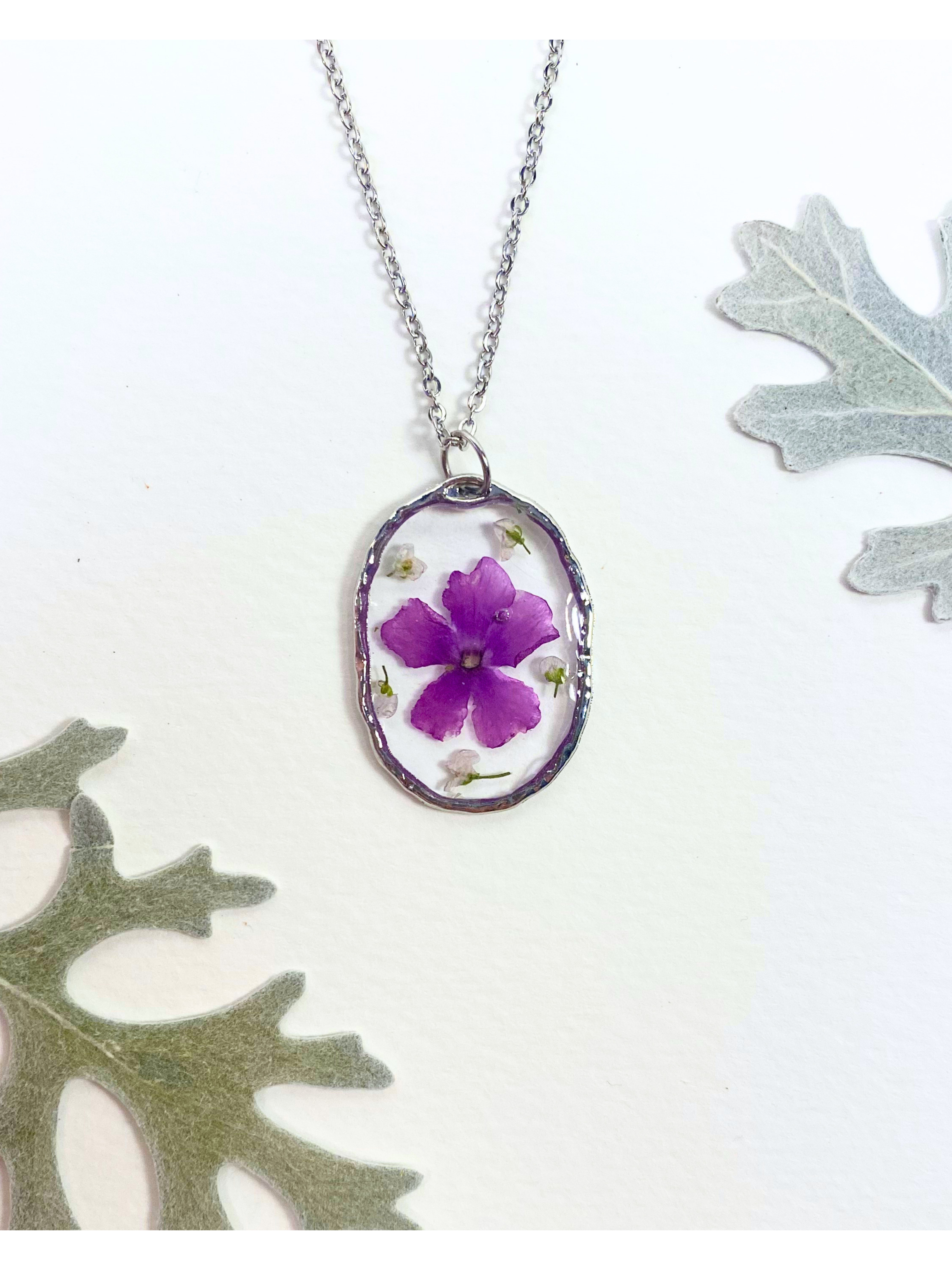 A silver oval pendant necklace featuring a preserved purple flower and small white flowers, displayed on a white background with decorative leaves.