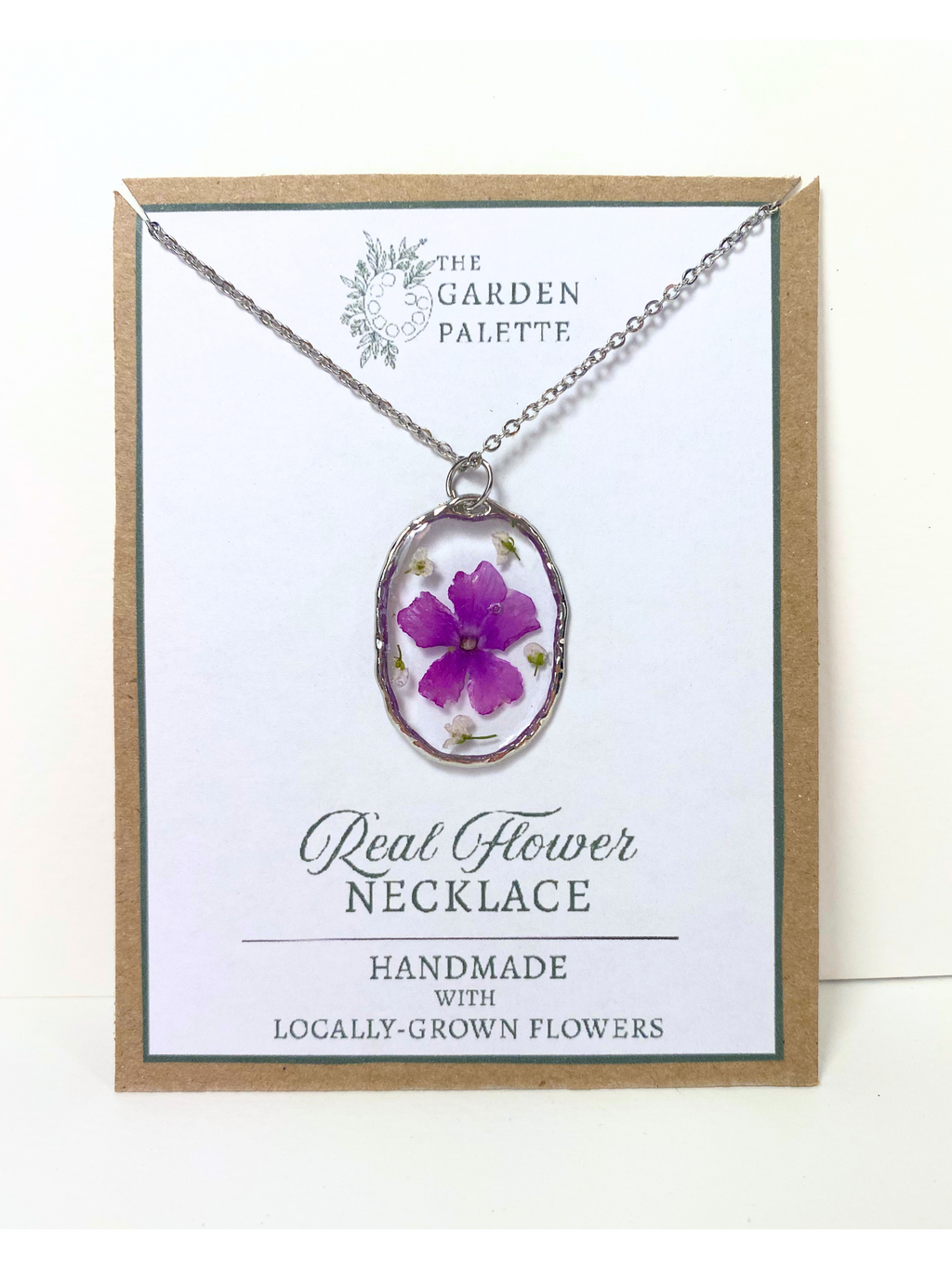 A handmade oval pendant necklace featuring a purple flower preserved in resin, displayed on a white card with a kraft border. The card is branded 'The Garden Palette' and reads 'Real Flower Necklace'.