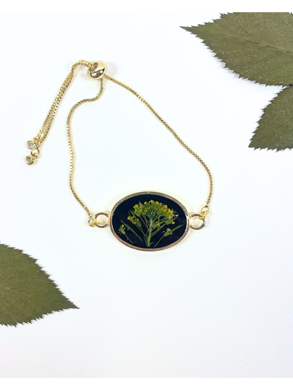 Gold chain bracelet with an oval pendant featuring a pressed yellow flower on a black background, styled with green leaves on a white surface.