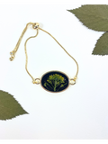 Gold chain bracelet with an oval pendant featuring a pressed yellow flower on a black background, styled with green leaves on a white surface.