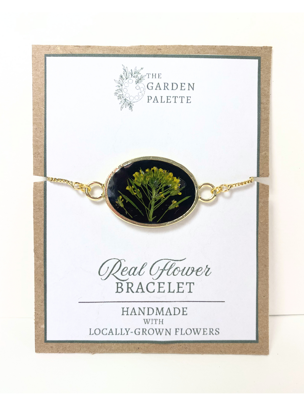 A gold-tone oval flower bracelet displayed on a branded card. The bracelet features a dark oval center with yellow real flowers, and the card reads 'The Garden Palette' and 'Real Flower Bracelet'.