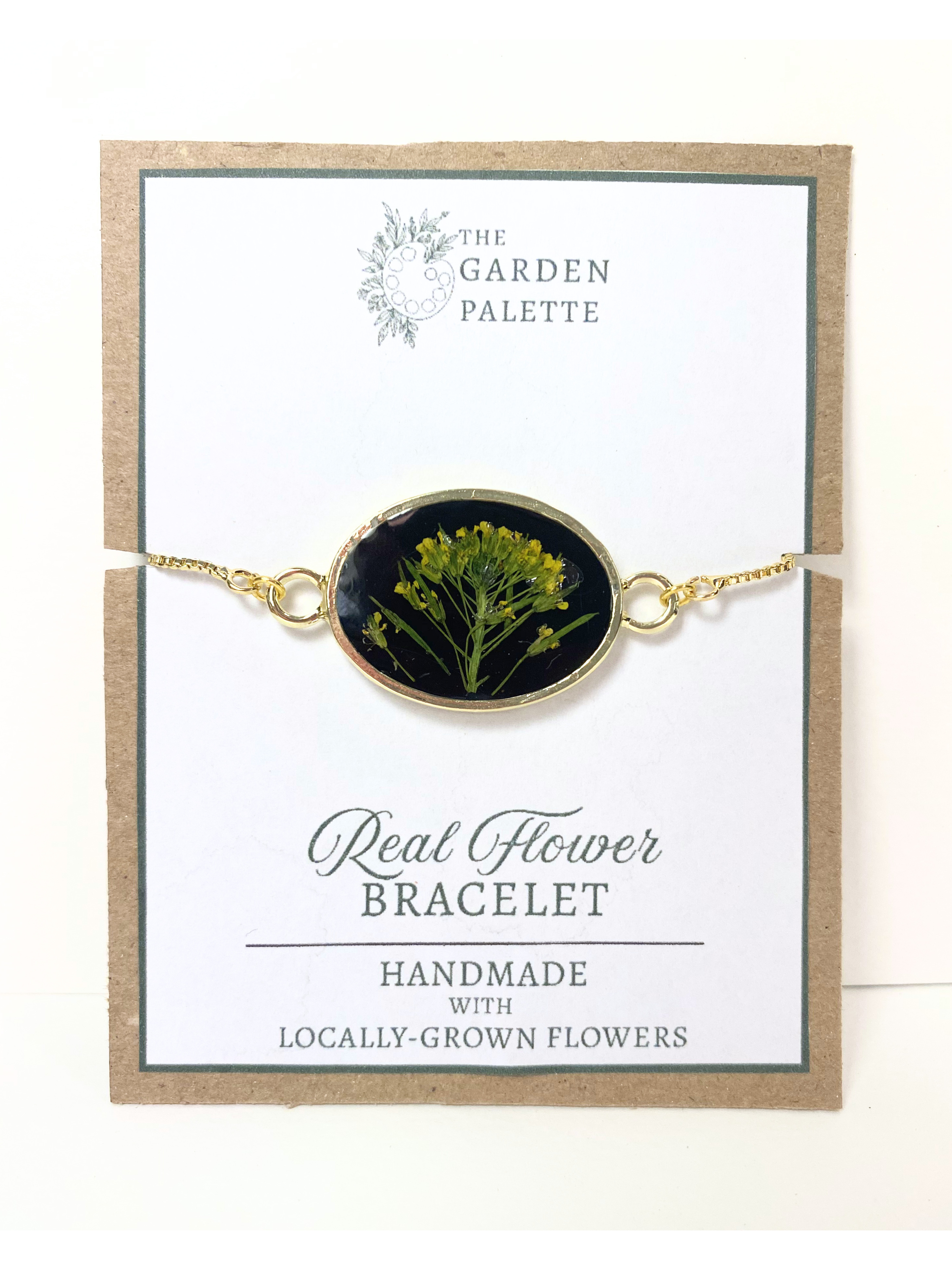 A gold-tone oval flower bracelet displayed on a branded card. The bracelet features a dark oval center with yellow real flowers, and the card reads 'The Garden Palette' and 'Real Flower Bracelet'.
