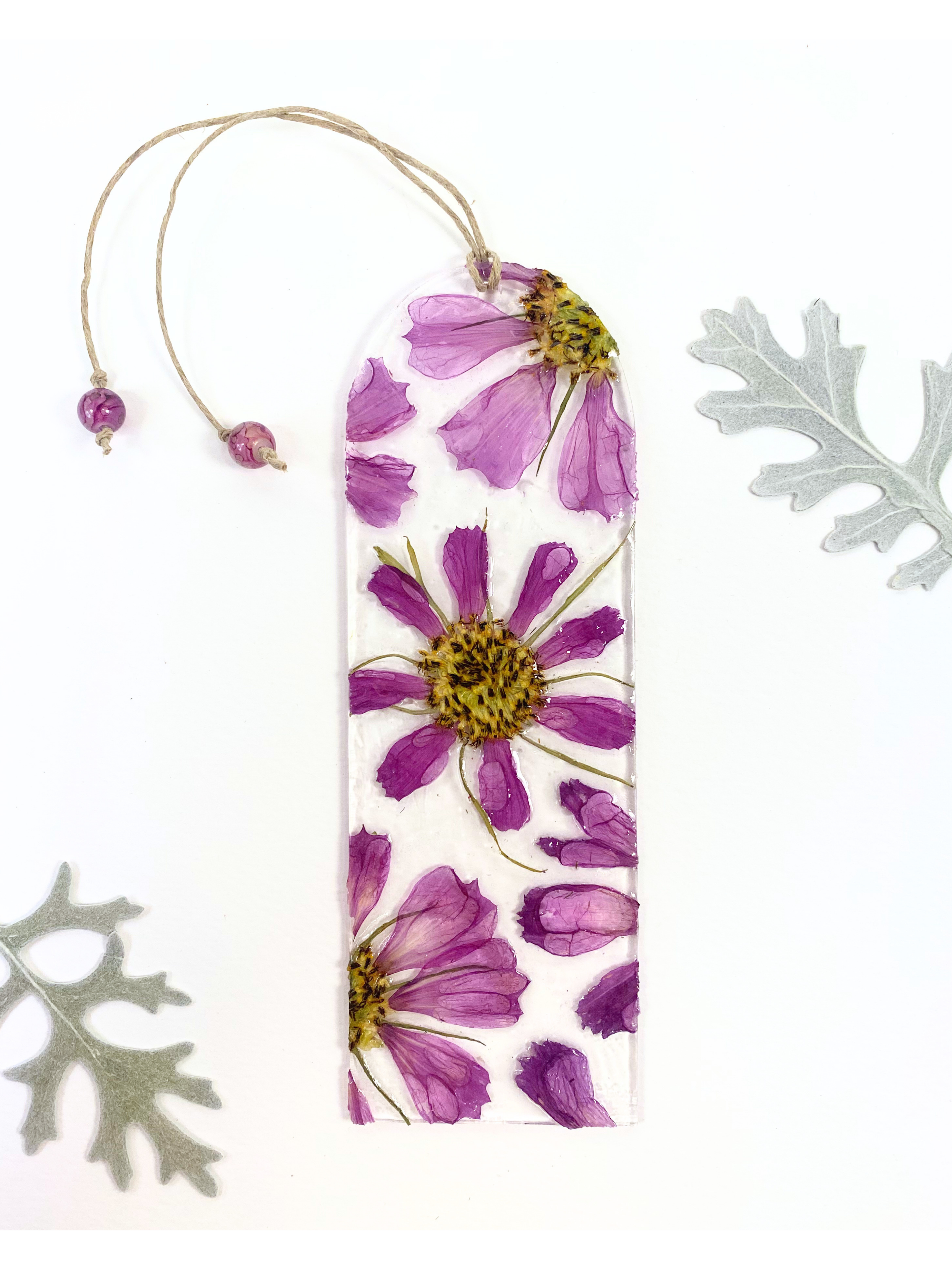 A bookmark featuring pressed purple flowers with yellow centers on a white background with decorative leaves at the corners.