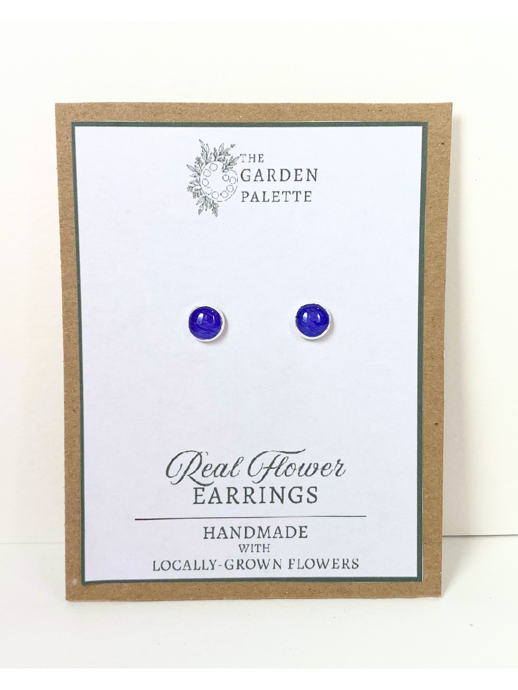 A pair of round silver stud earrings containing deep blue flower petals, mounted on a white display card with 'The Garden Palette' branding and 'Real Flower Earrings' text.