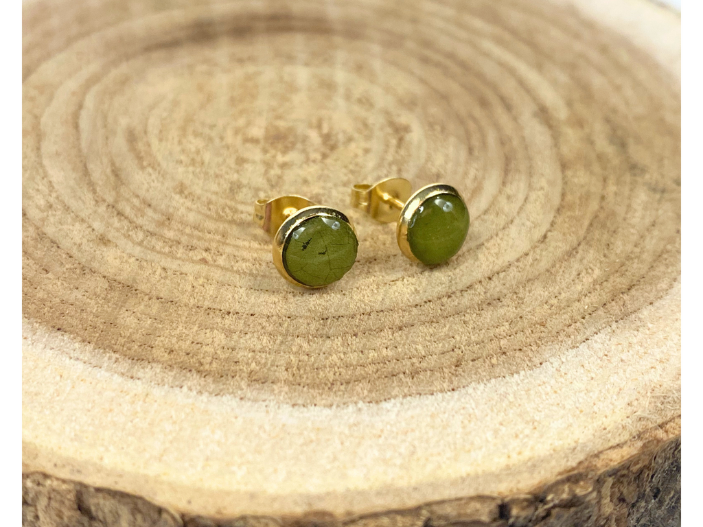 A pair of round gold stud earrings containing green leaves, displayed on a rustic wood slice background.