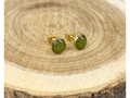 A pair of round gold stud earrings containing green leaves, displayed on a rustic wood slice background.
