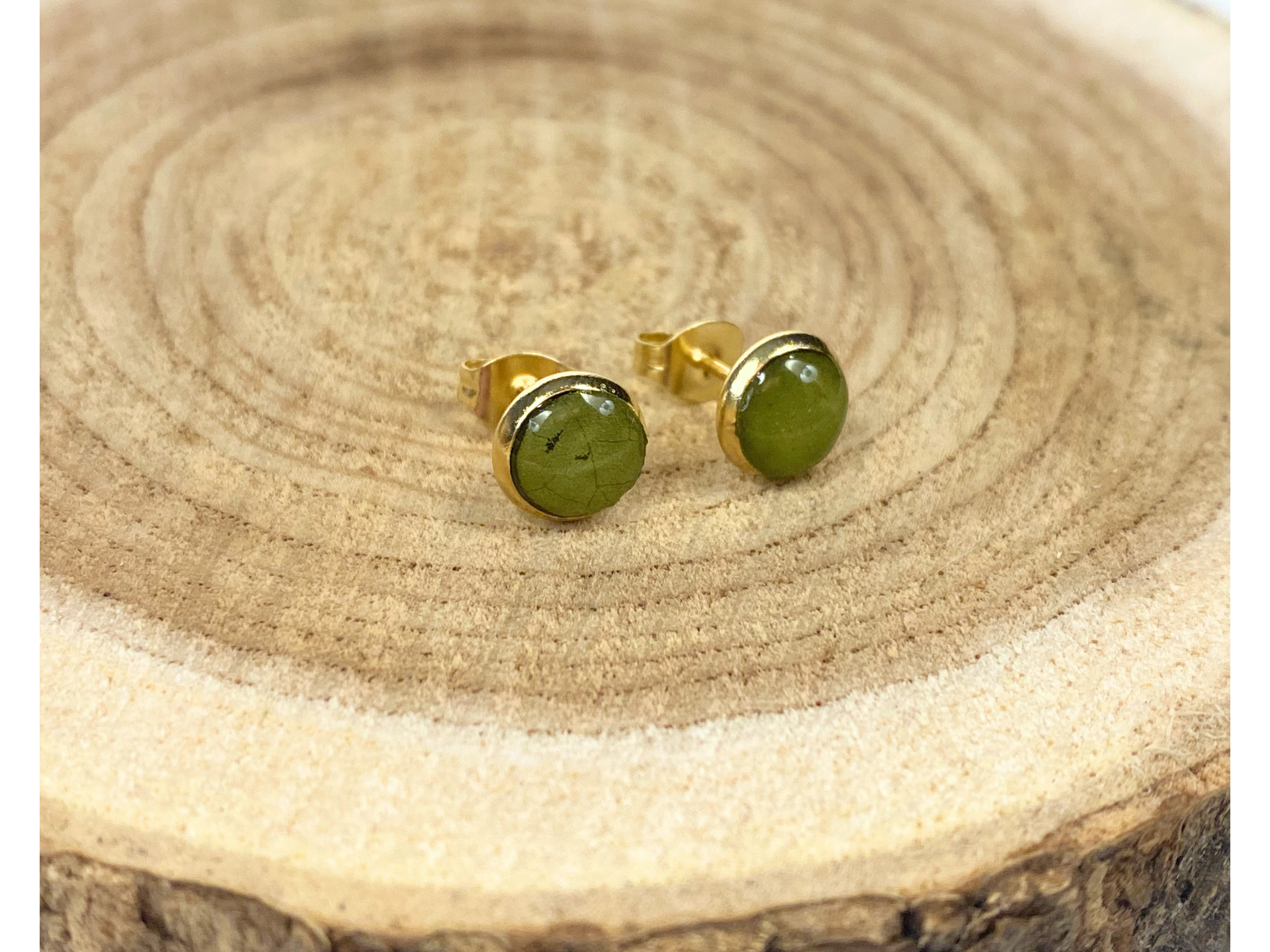 A pair of round gold stud earrings containing green leaves, displayed on a rustic wood slice background.