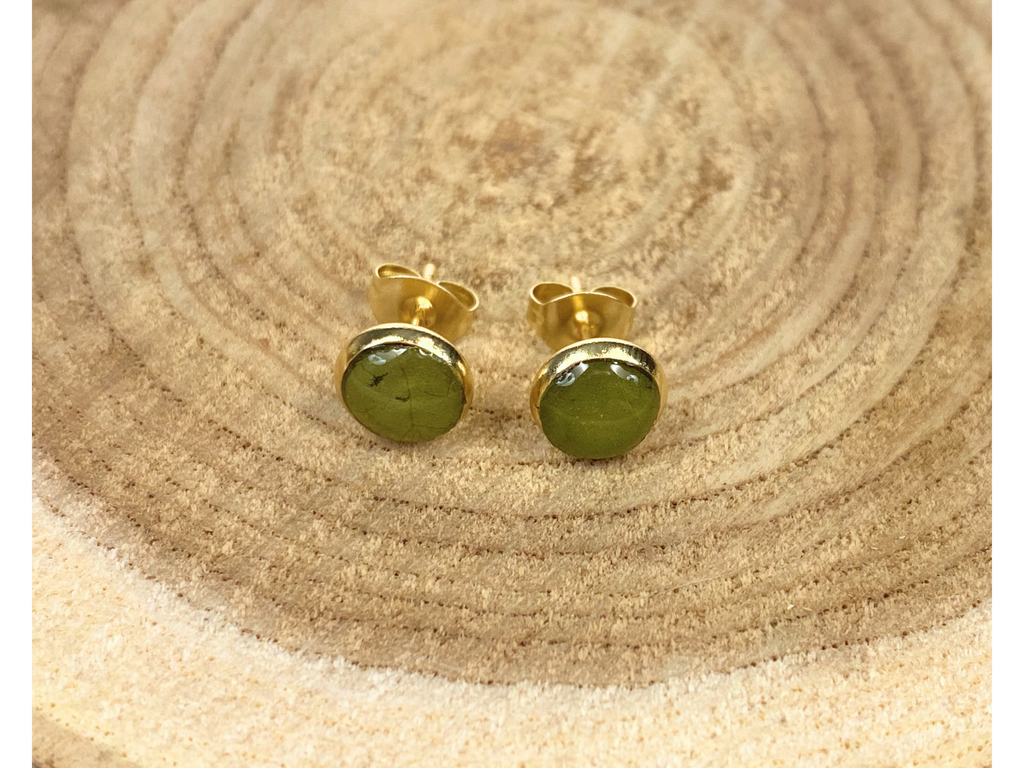 A pair of round gold stud earrings containing green leaves, displayed on a rustic wood slice background.