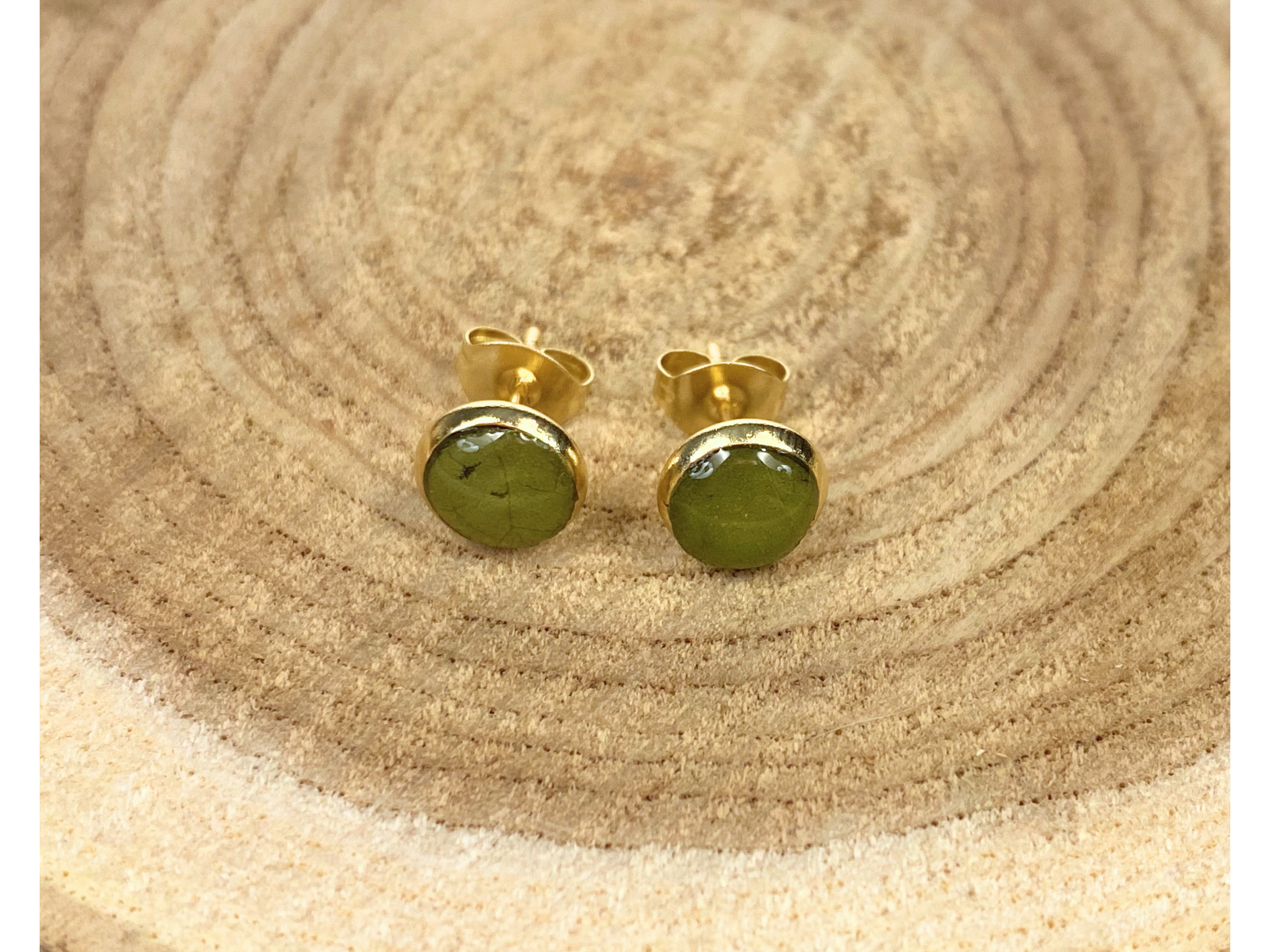 A pair of round gold stud earrings containing green leaves, displayed on a rustic wood slice background.