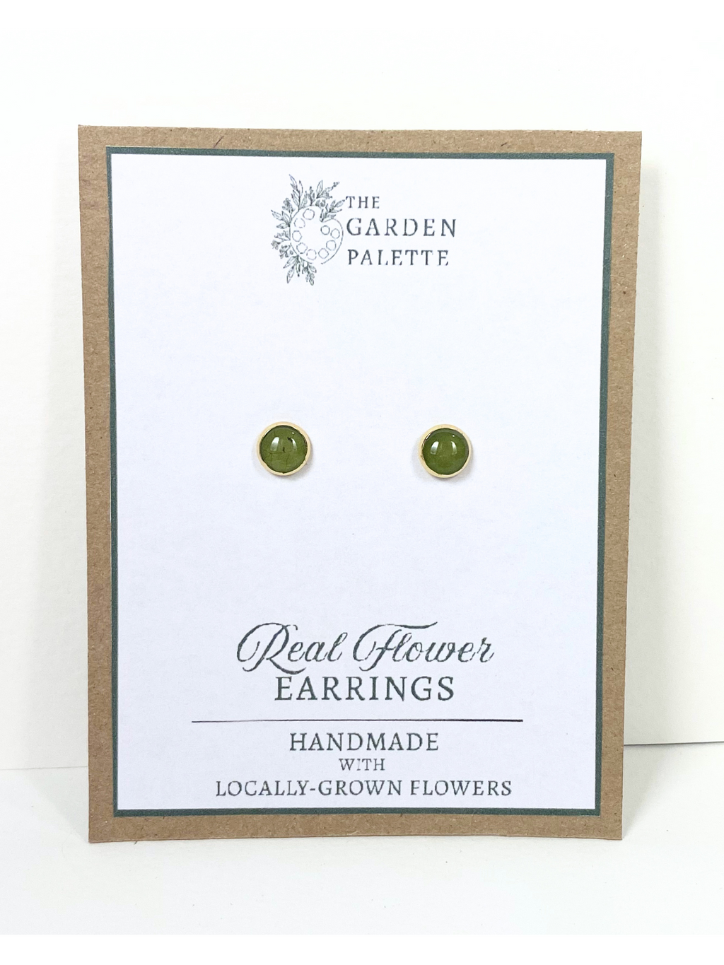 A pair of small round stud earrings containing gtreen leaves mounted on a white display card with a kraft border. The card reads 'The Garden Palette' and 'Real Flower Earrings'.