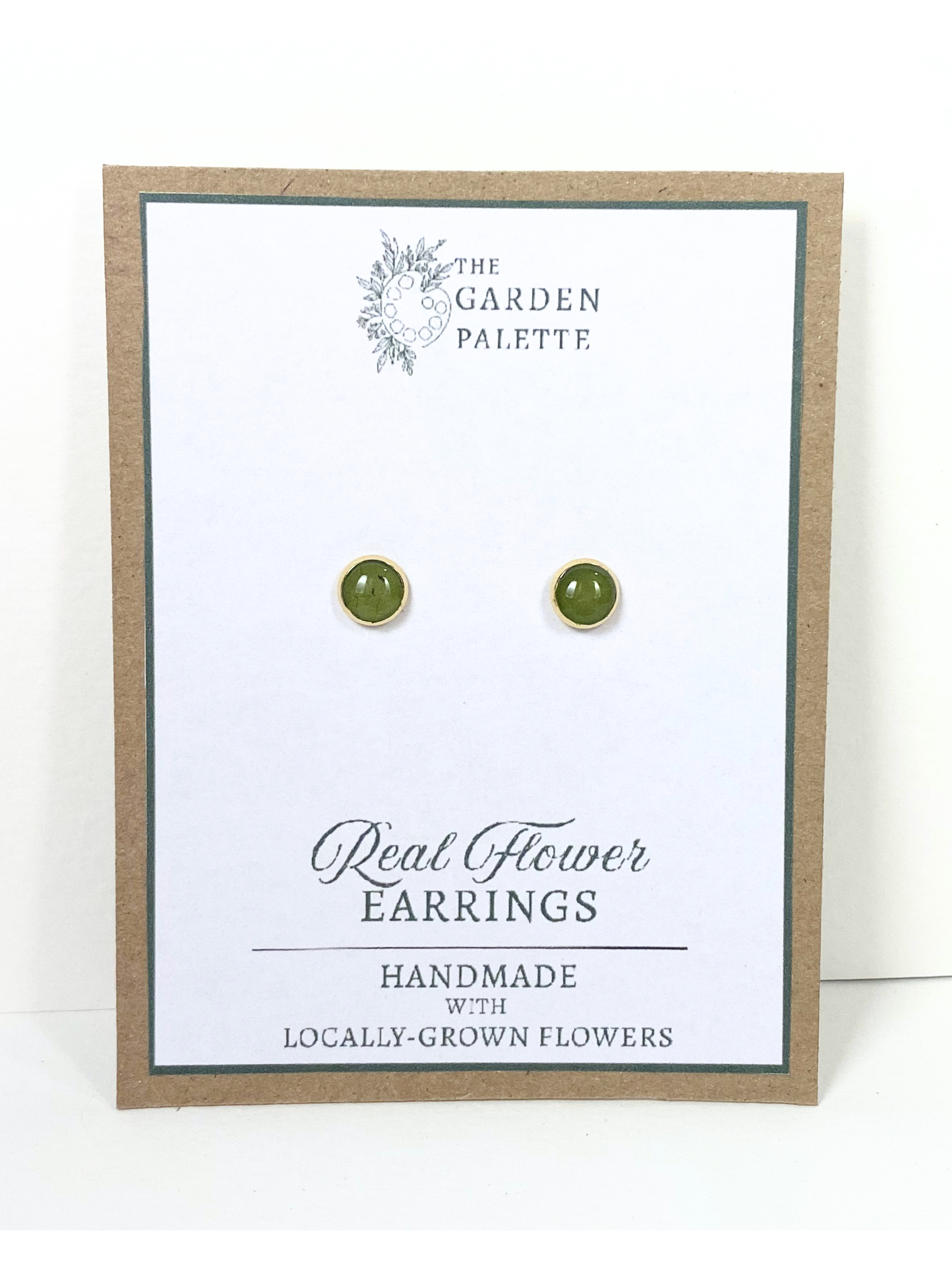 A pair of small round stud earrings containing gtreen leaves mounted on a white display card with a kraft border. The card reads 'The Garden Palette' and 'Real Flower Earrings'.